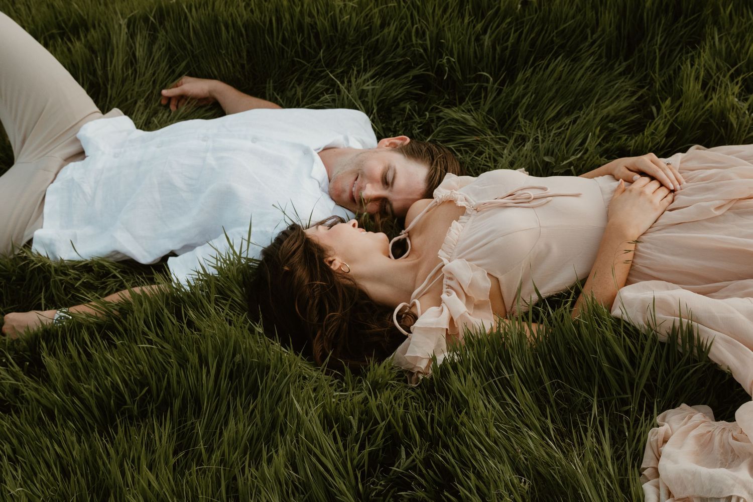 Two people in light flowy outfits lie close together in tall grass on a warm summer evening.