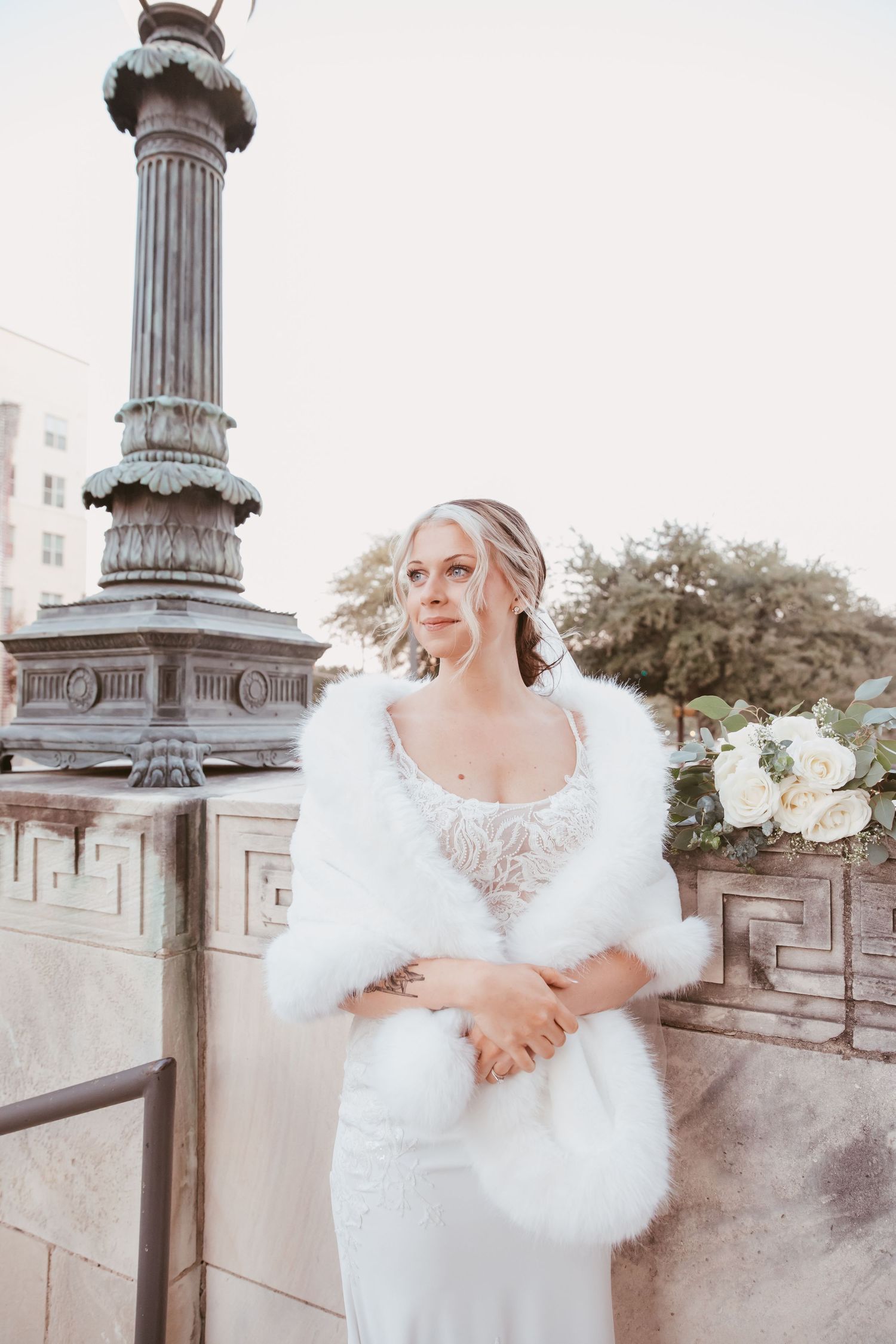 Elegant bride in white lace gown and fur shawl poses gracefully by ornate stone pillars in winter wedding portraits.