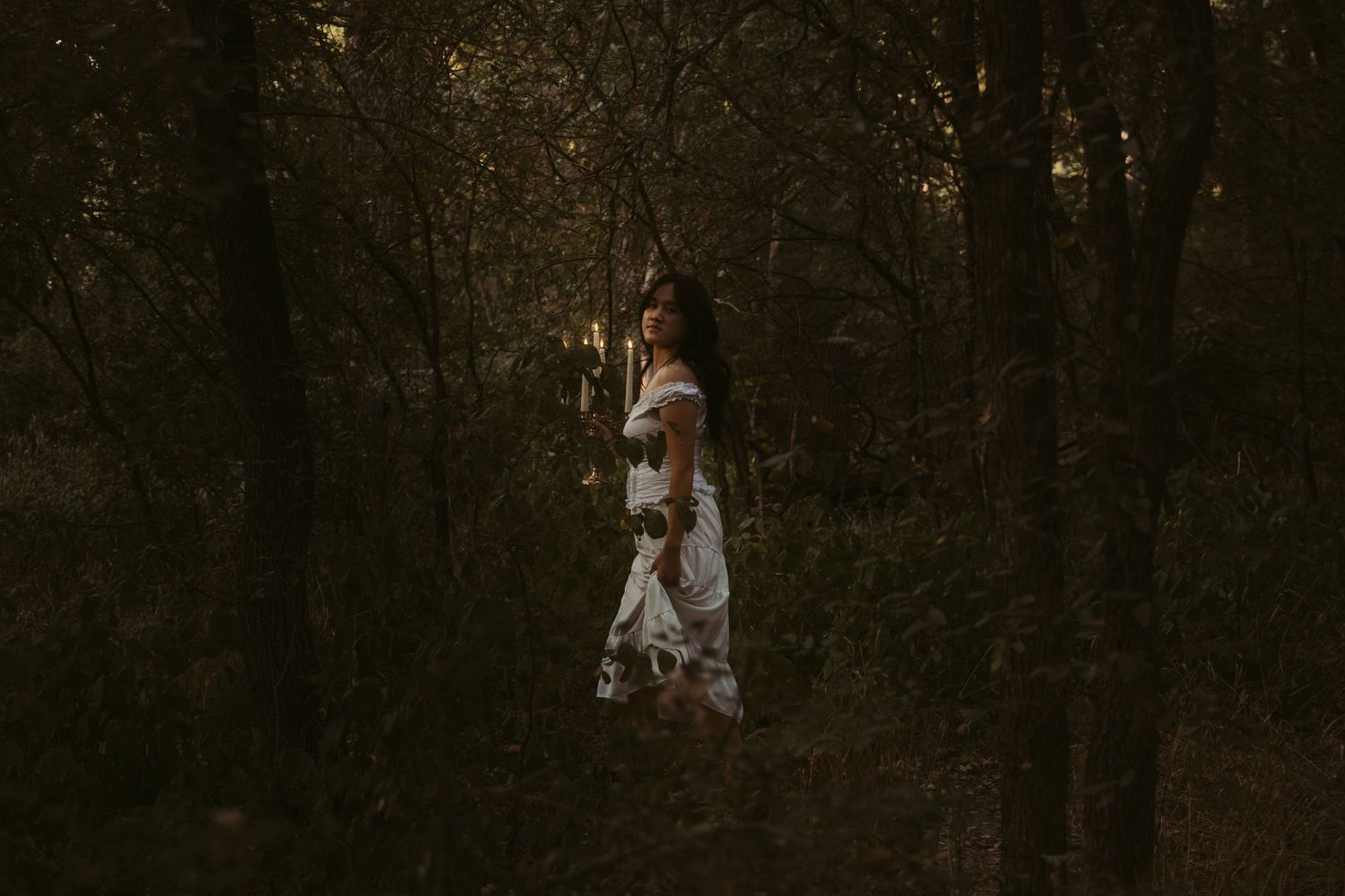 A person in a white dress stands in a dark forest as sunlight filters through the trees creating a moody atmosphere.