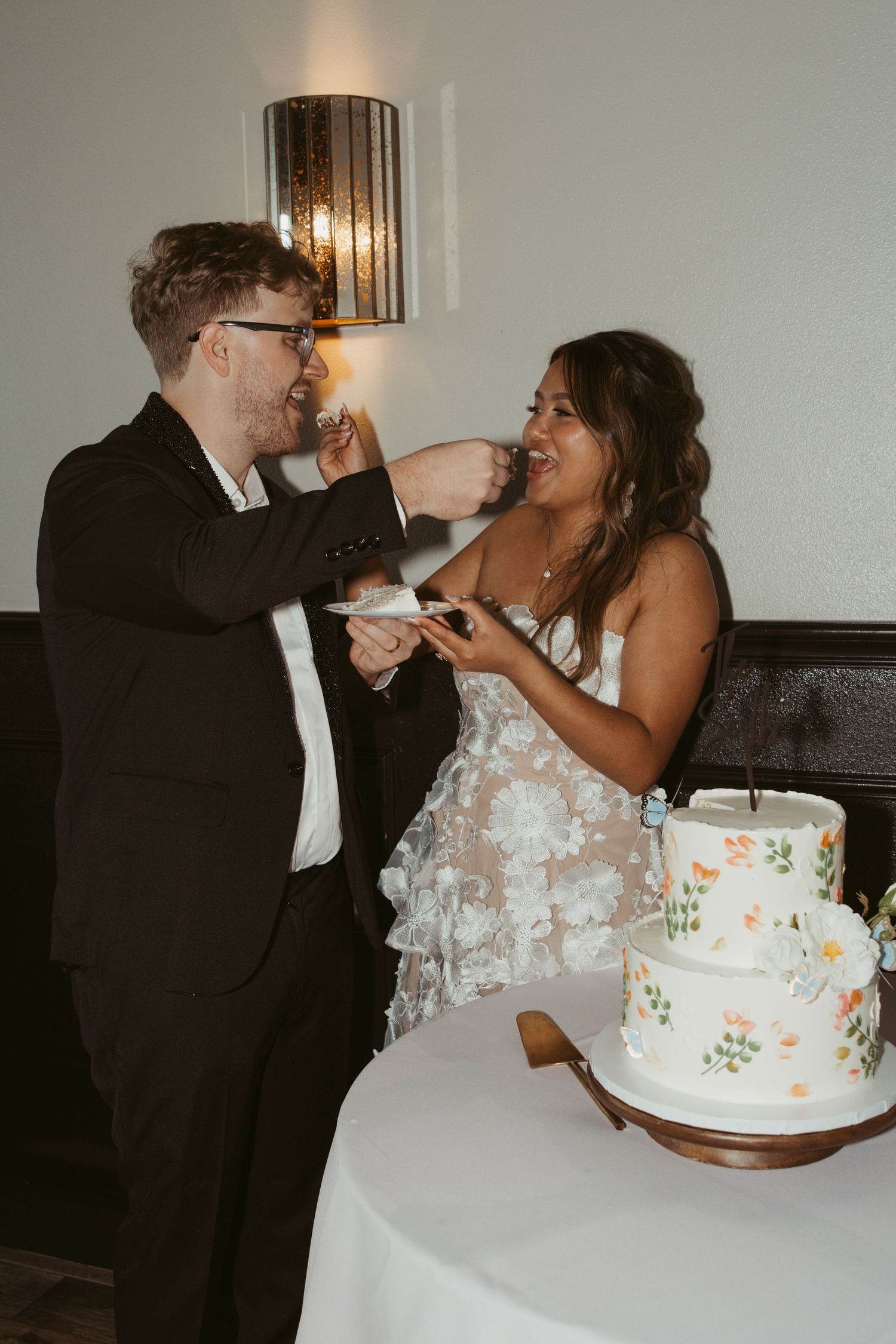 Romantic cake cutting moment at wedding reception with tiered white cake decorated with delicate floral details.