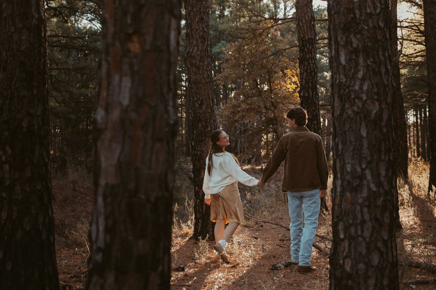 A couple walks hand in hand through a sunlit autumn forest with tall pine trees surrounding them.