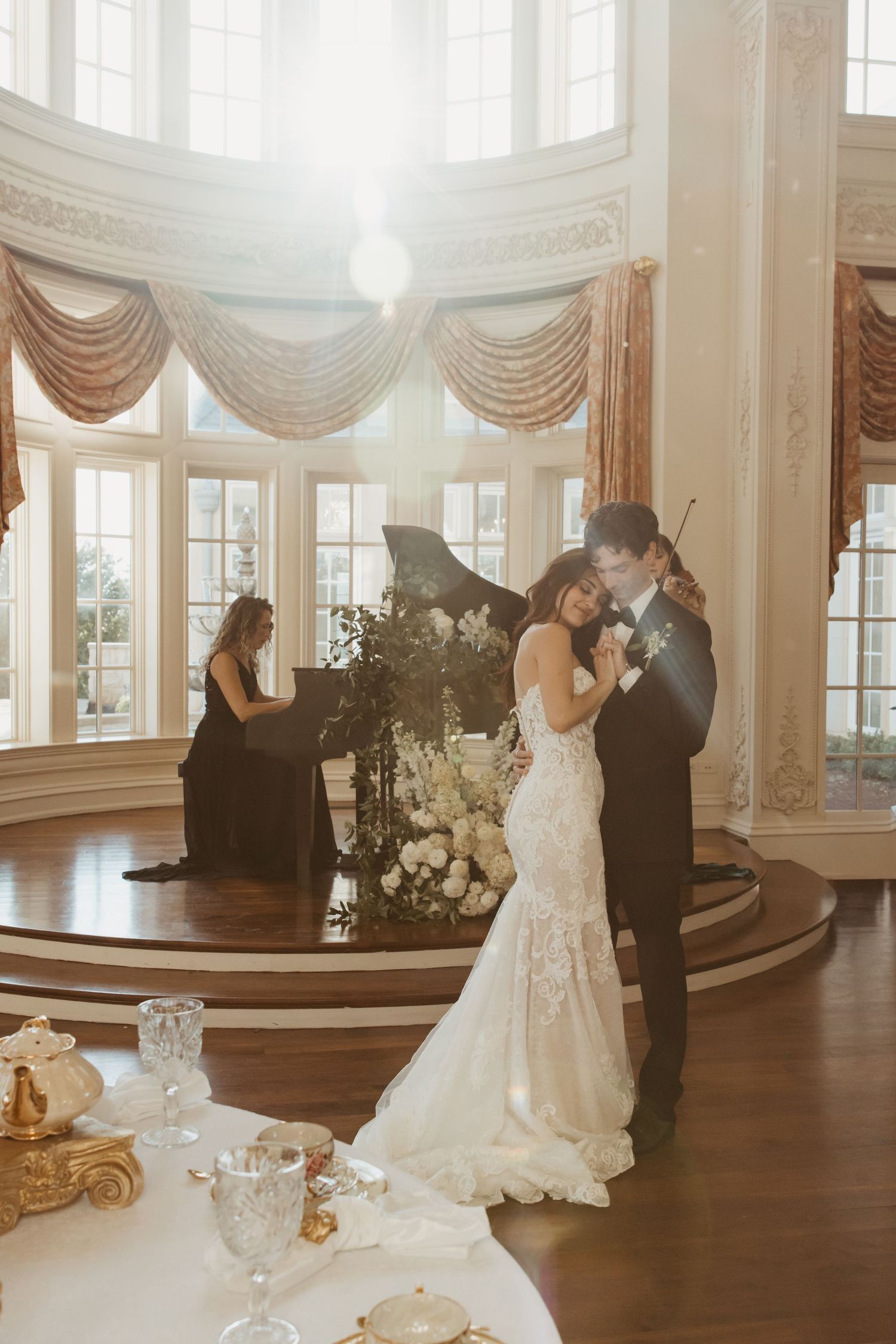 Intimate wedding moment in sunlit ballroom with piano and elegant table settings.