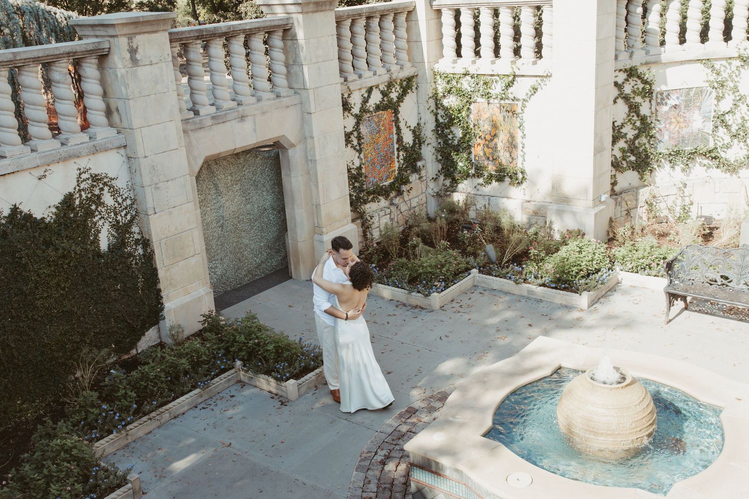 A couple embraces in an elegant garden courtyard with white stone balustrades and a decorative fountain. Wedding photographer DFW