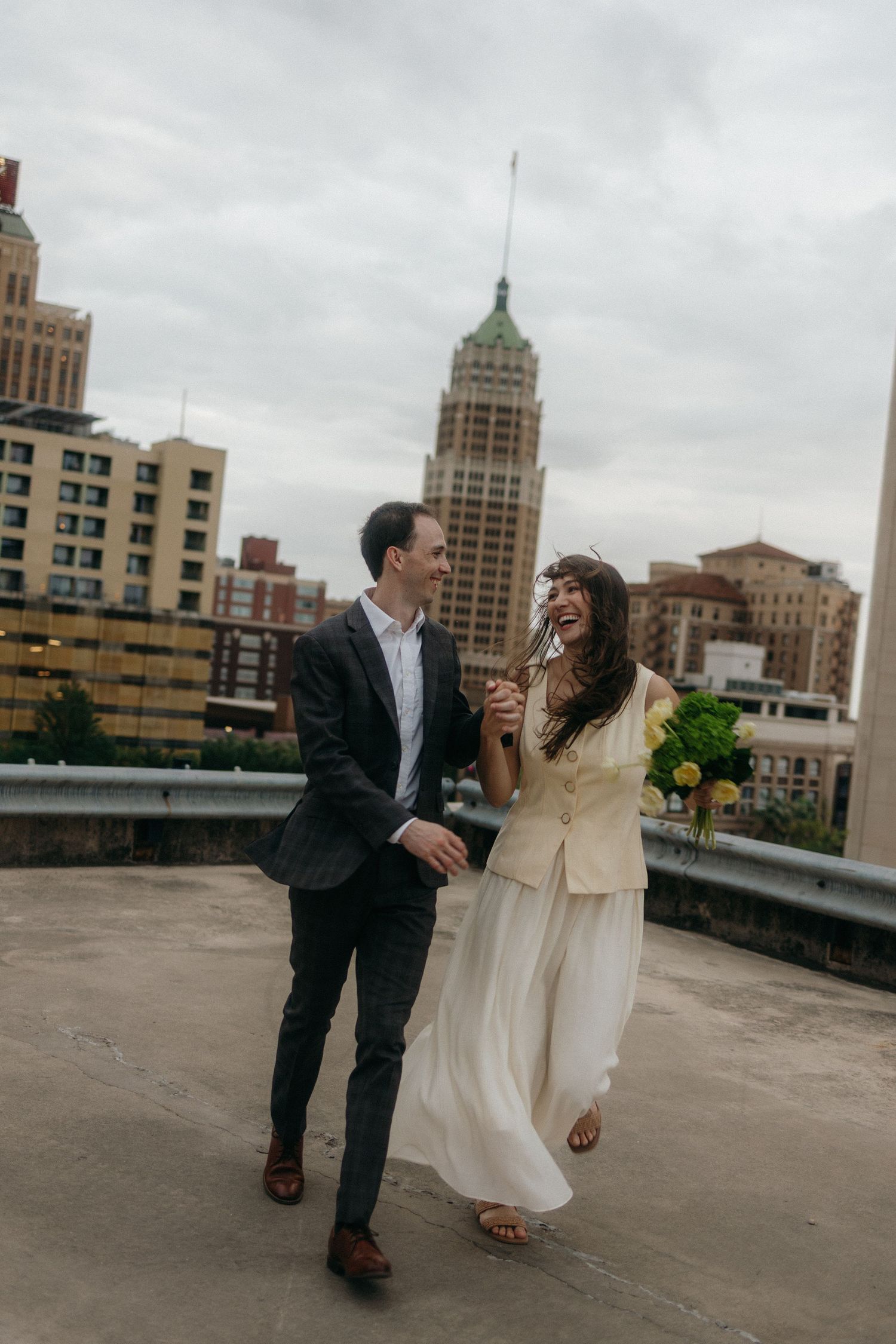 Couple laughing and walking on rooftop with city skyline backdrop on their wedding day.