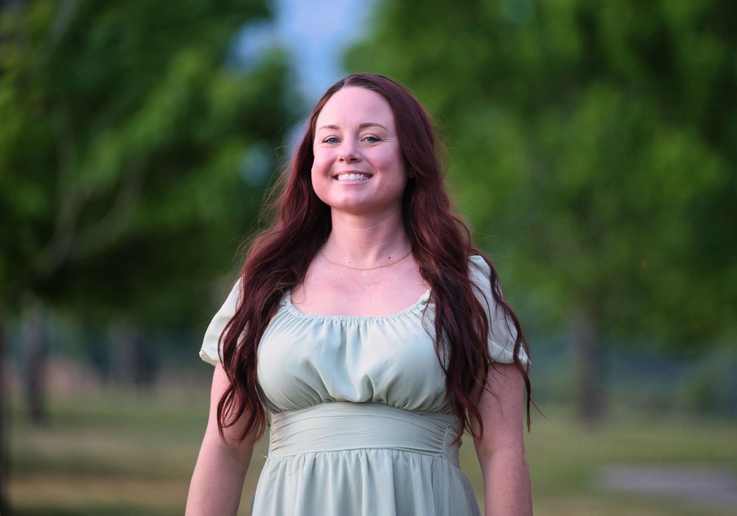 Smiling woman with long red hair wearing a sage green dress standing in a lush green park.