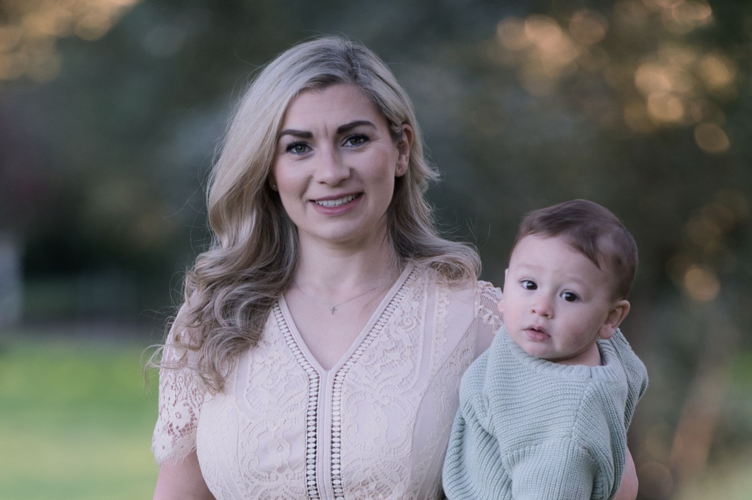 A smiling blonde woman in a pink lace dress holds a baby outdoors in a park setting.