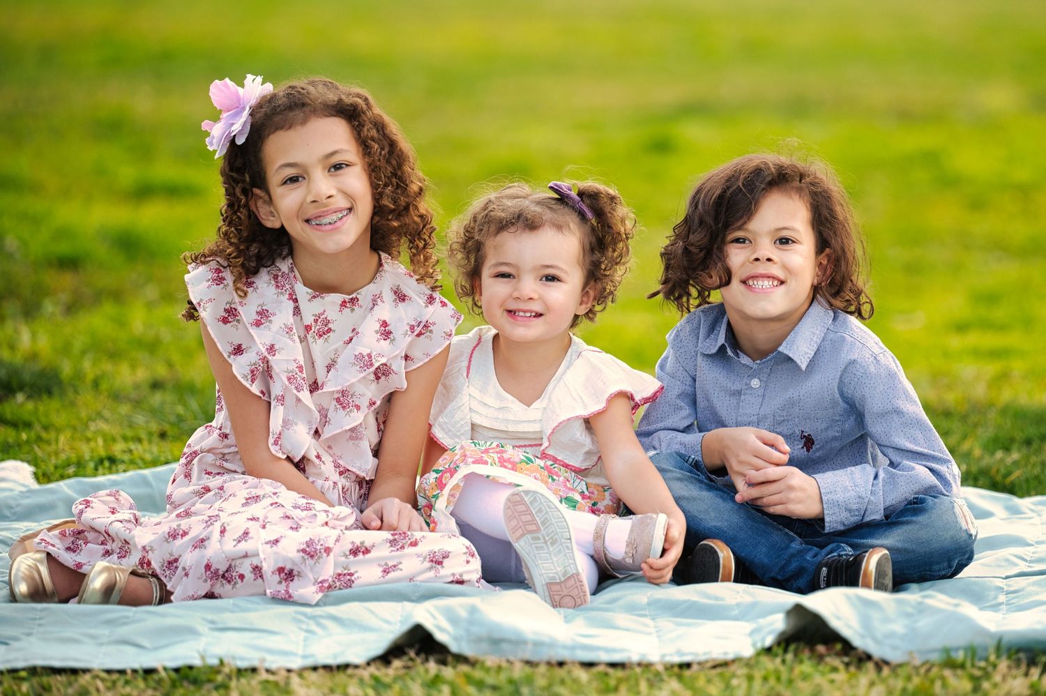 Three young children sitting on blanket in grass, smiling for family portrait outdoors.