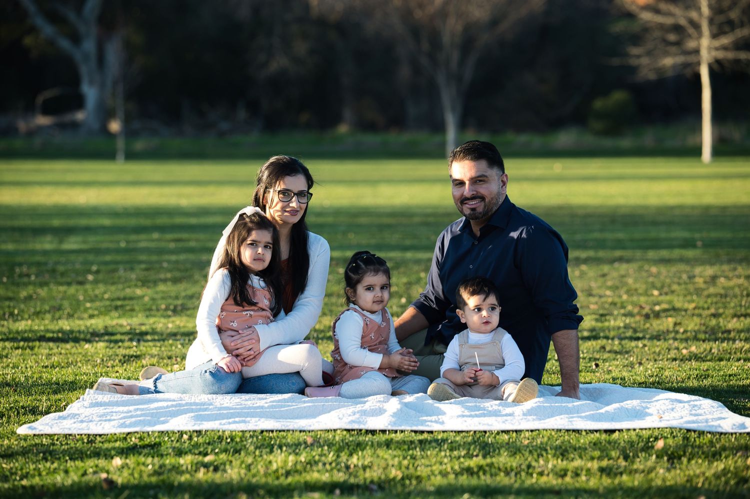 A family of five sits together on a white blanket in a sunny green park for a portrait.