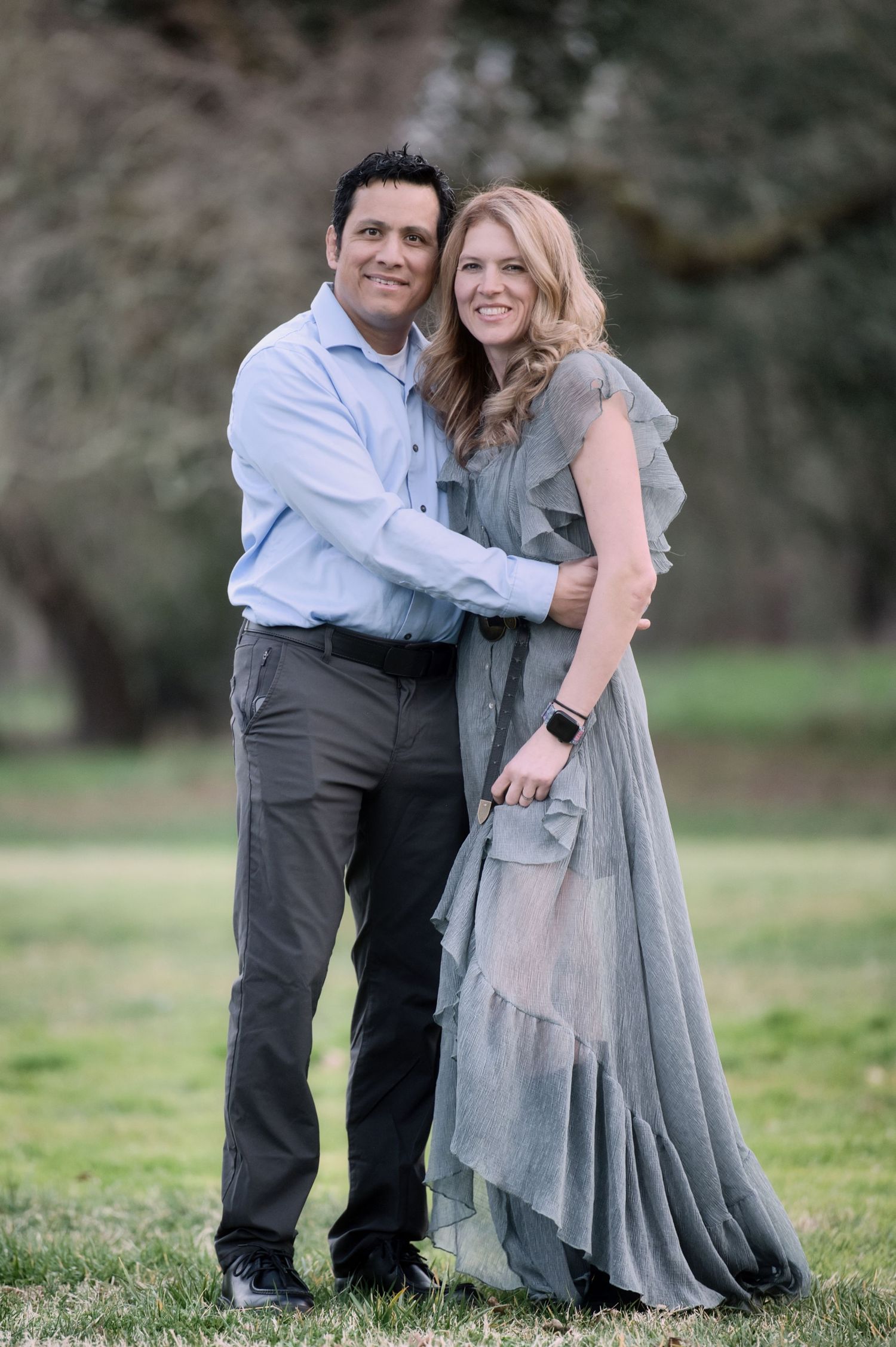 A smiling couple embraces outdoors in a park, dressed in elegant gray and blue attire.