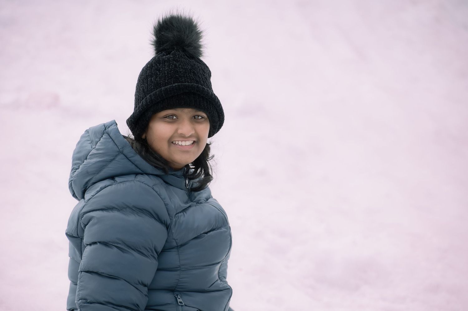 A young girl smiles in a gray puffer jacket and black pom-pom beanie against a snowy background.