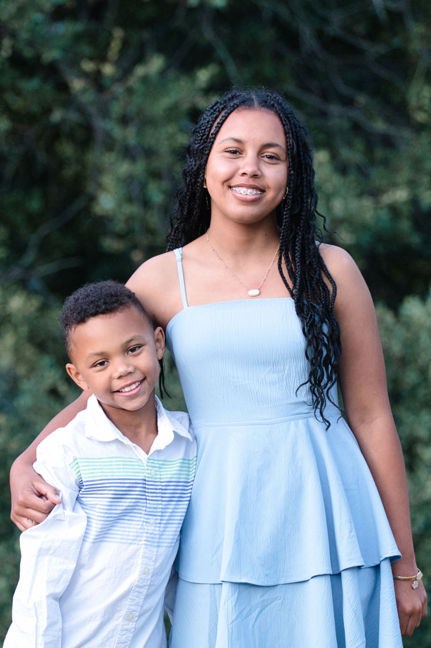 Smiling teenage girl in blue dress poses with young boy in white shirt in an outdoor park portrait.