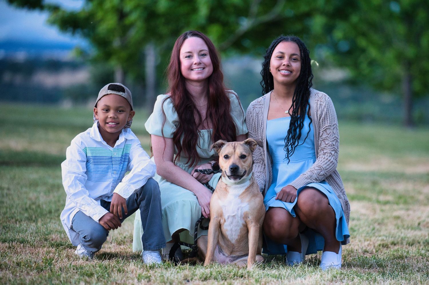 Three children posing with a tan dog on green grass in a park at dusk.