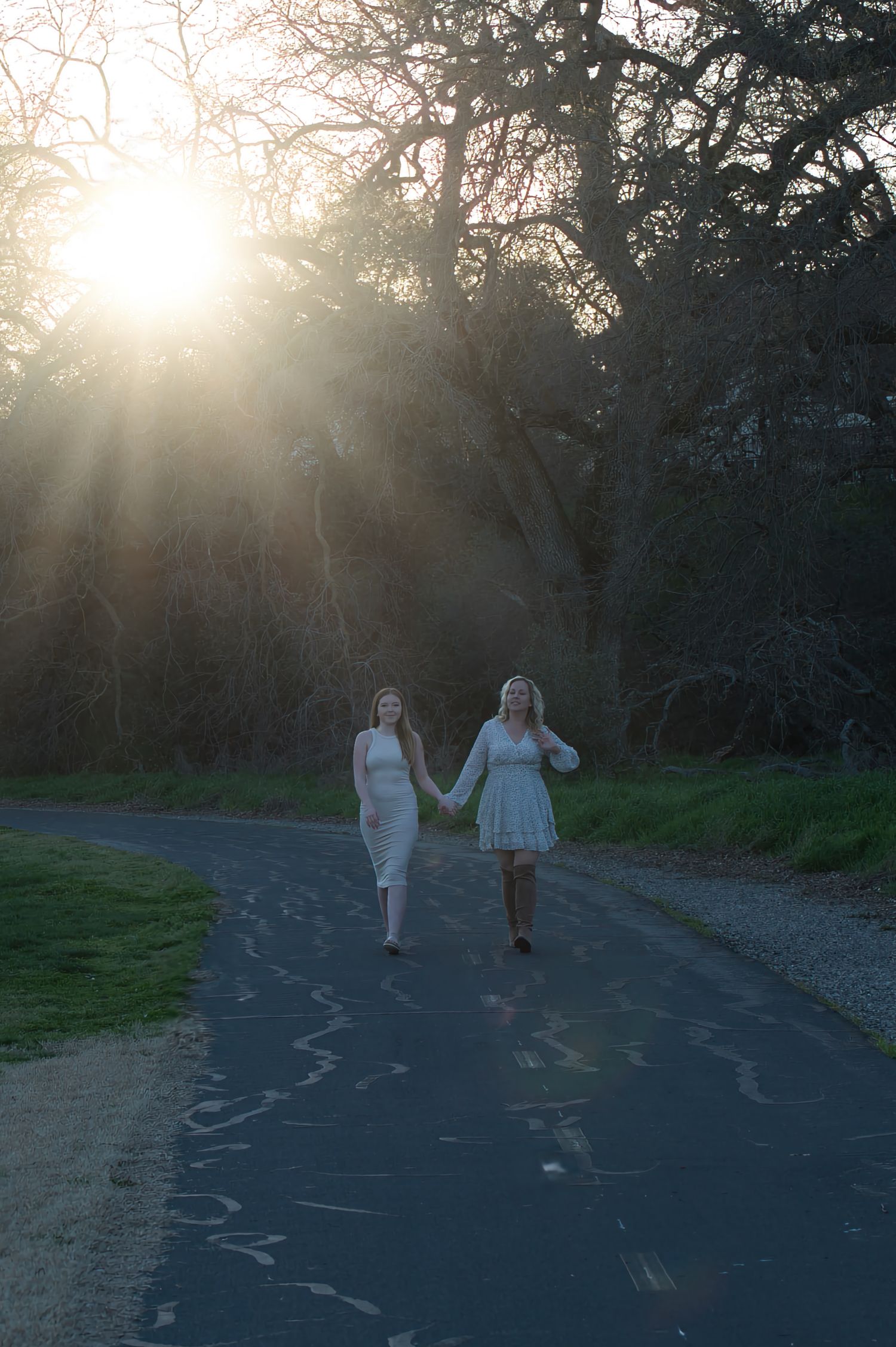 Two women in white dresses hold hands walking down a tree-lined road bathed in golden sunlight.