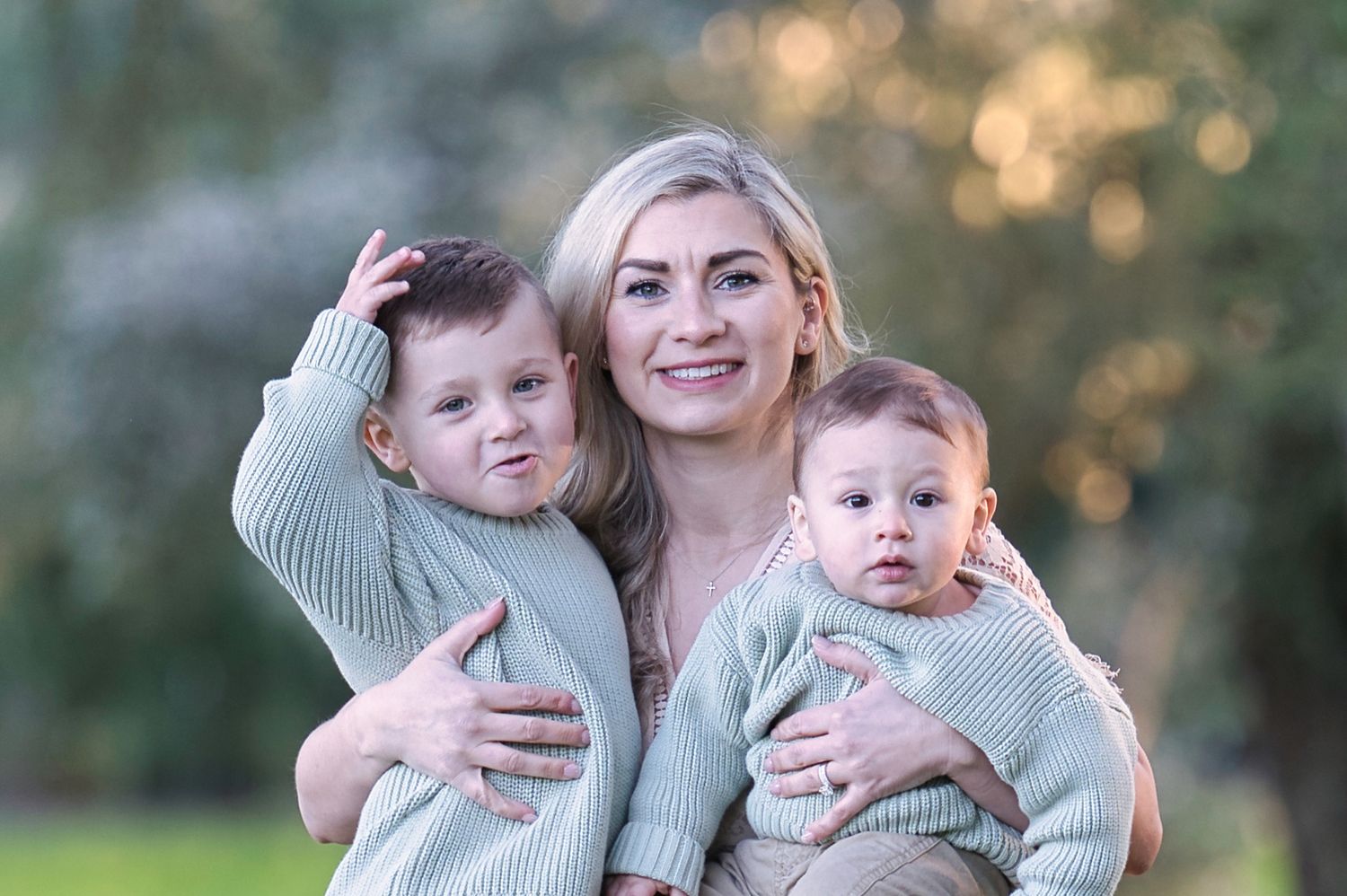 A smiling blonde mother cuddles two young boys in matching sage green sweaters outdoors.