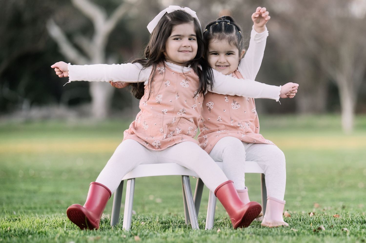 Two little girls in matching pink outfits and red boots pose playfully on chairs outdoors.