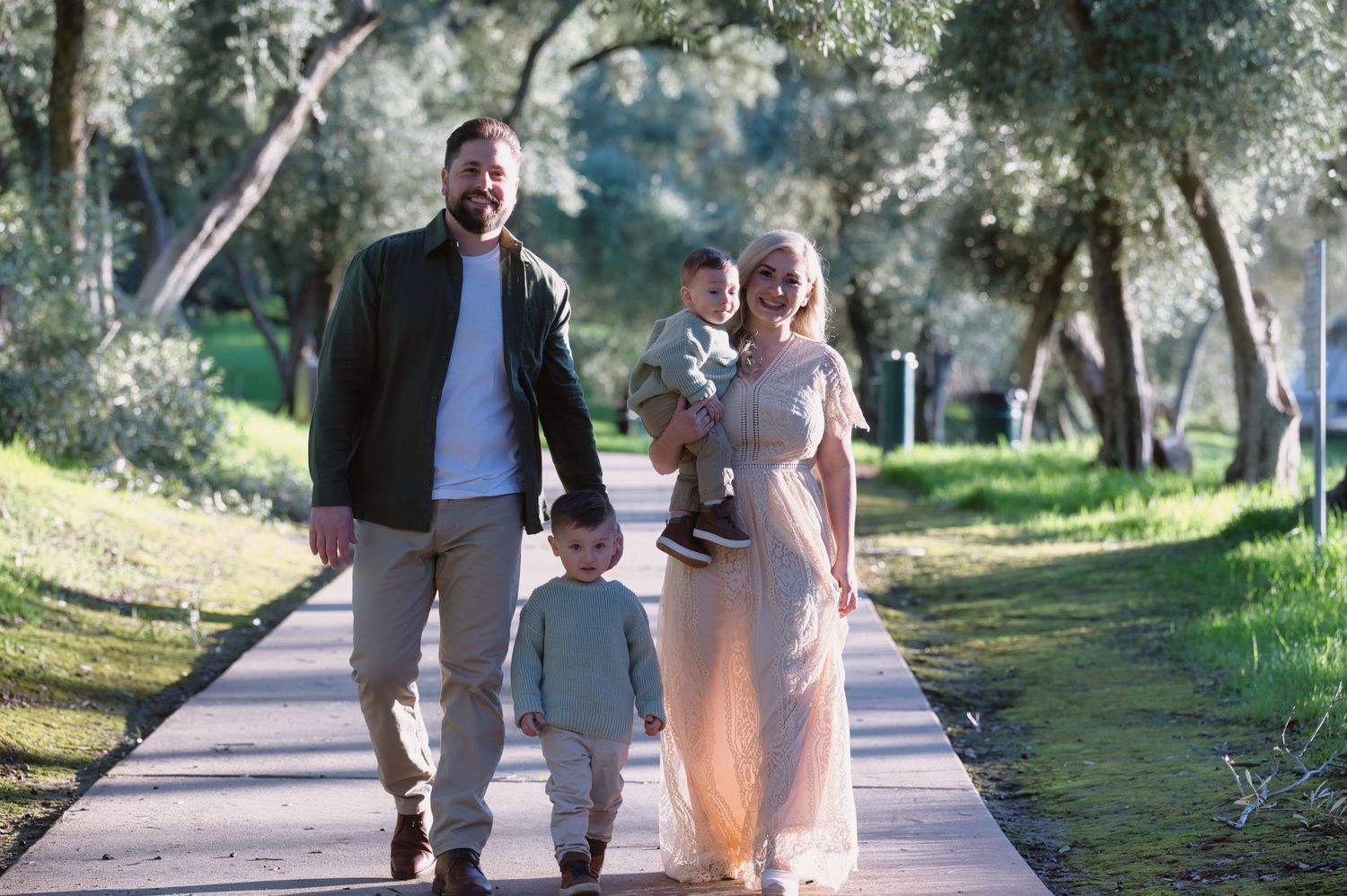 Young family of four walking along a tree-lined path in a sunny park, parents holding two small children.
