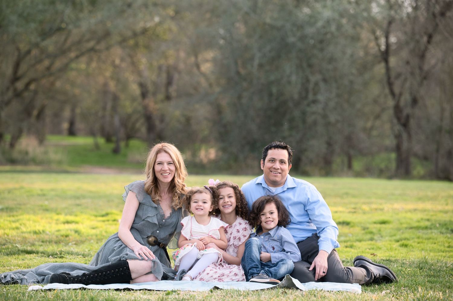 Family of five sitting on a blanket in a sunny park surrounded by blooming yellow wildflowers.
