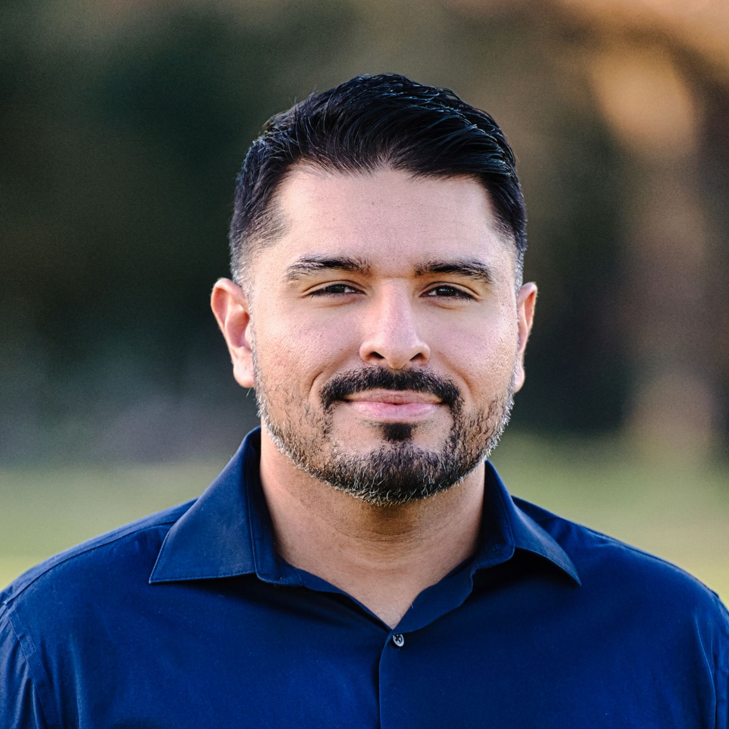 Smiling man with beard wearing navy blue shirt outdoors with blurred green background.