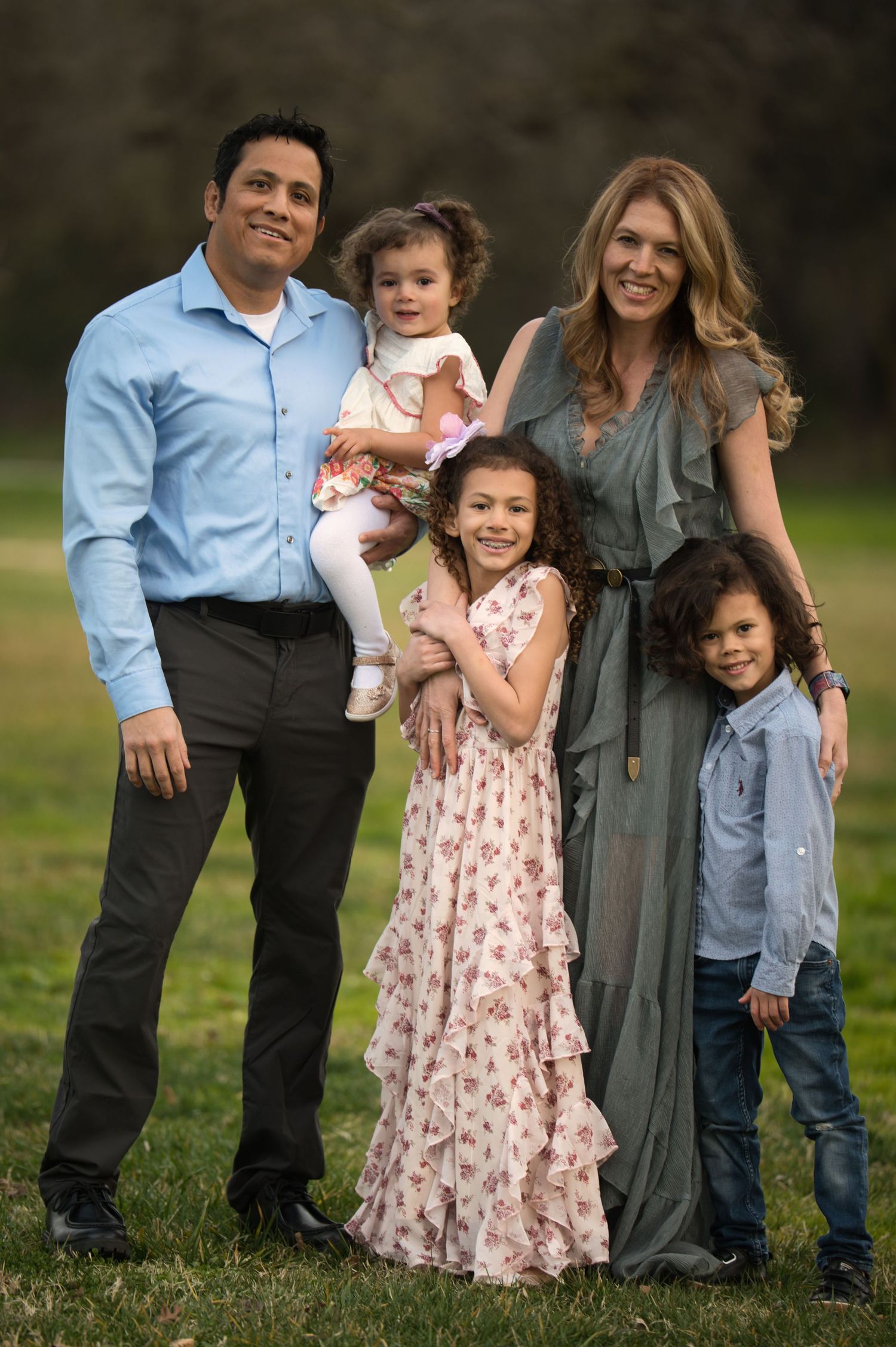 Smiling Hispanic family of five poses together in green outdoor park setting.