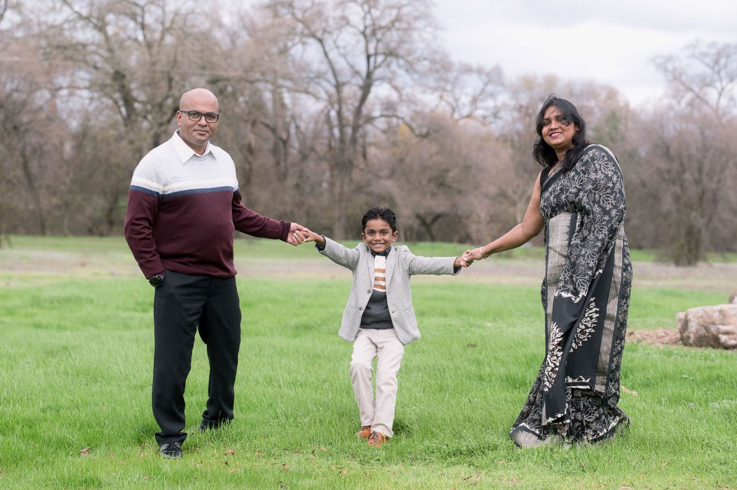 Family of three holding hands in a green park, parents and young boy smiling together.