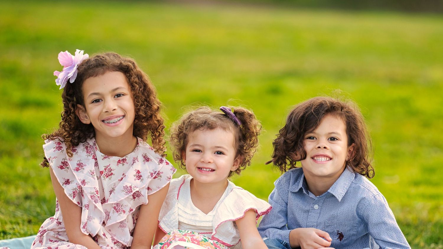 Three young siblings sit together smiling on a green lawn during an outdoor family photo session.