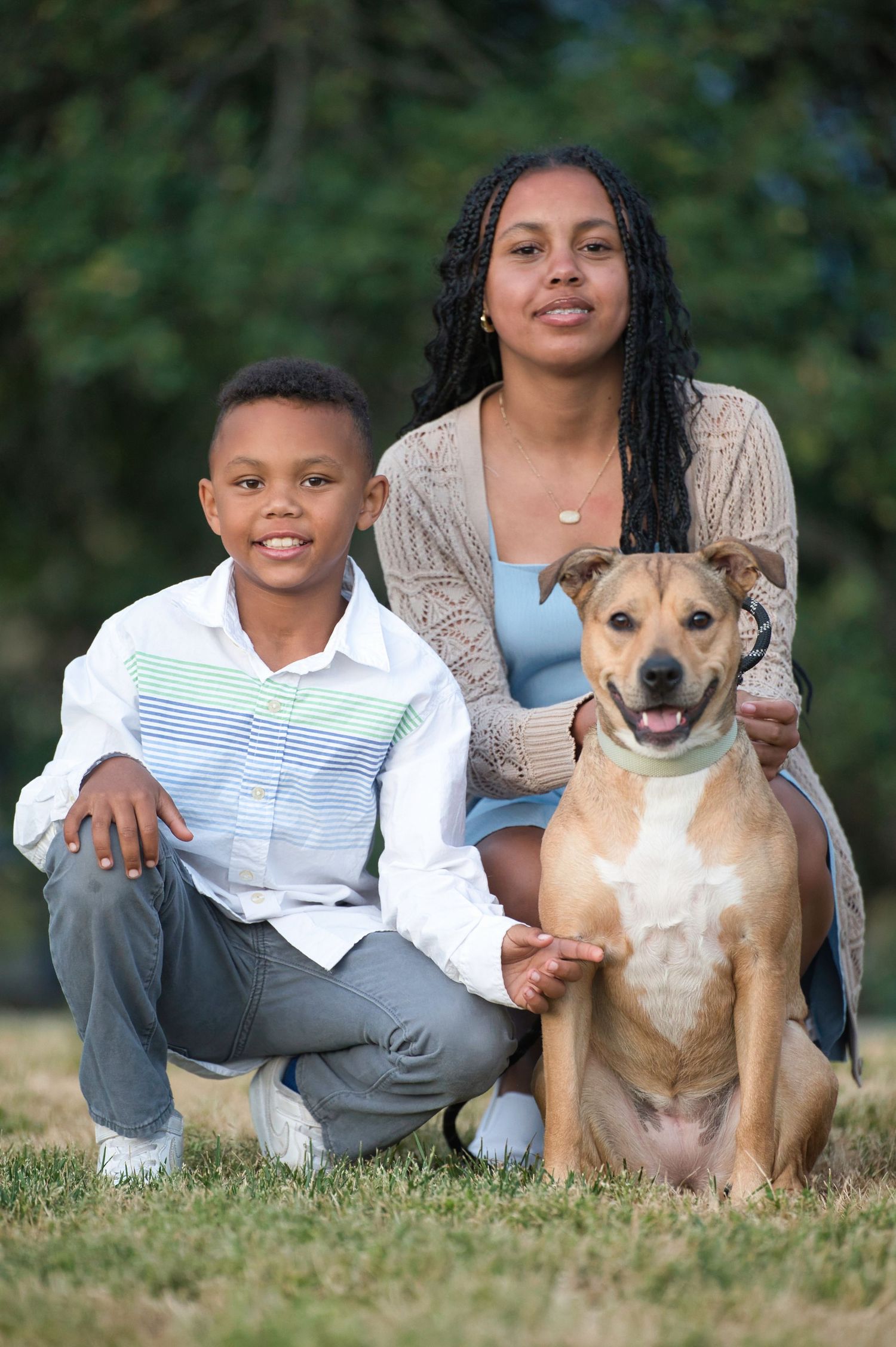 A woman and young boy smile with their tan dog in a lush green park setting.