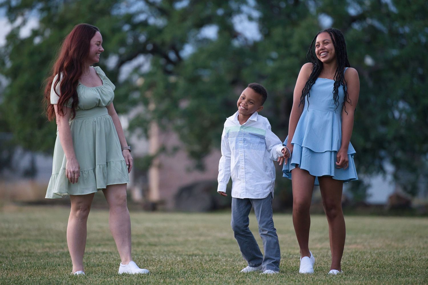 Two women in pastel dresses holding hands with a young boy smiling in a green park.