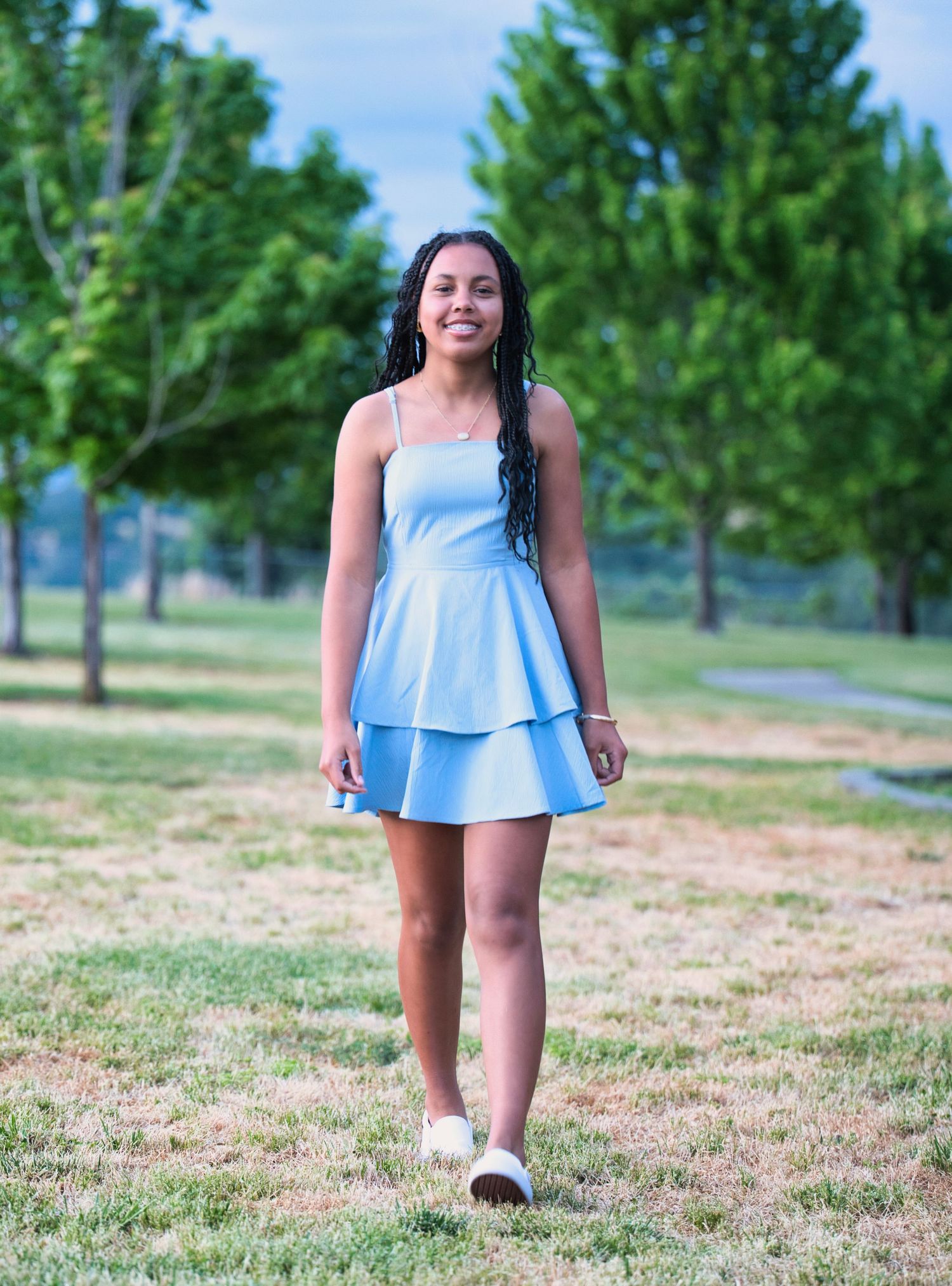 Young woman in light blue tiered mini dress walking outdoors in a green park on a sunny day.