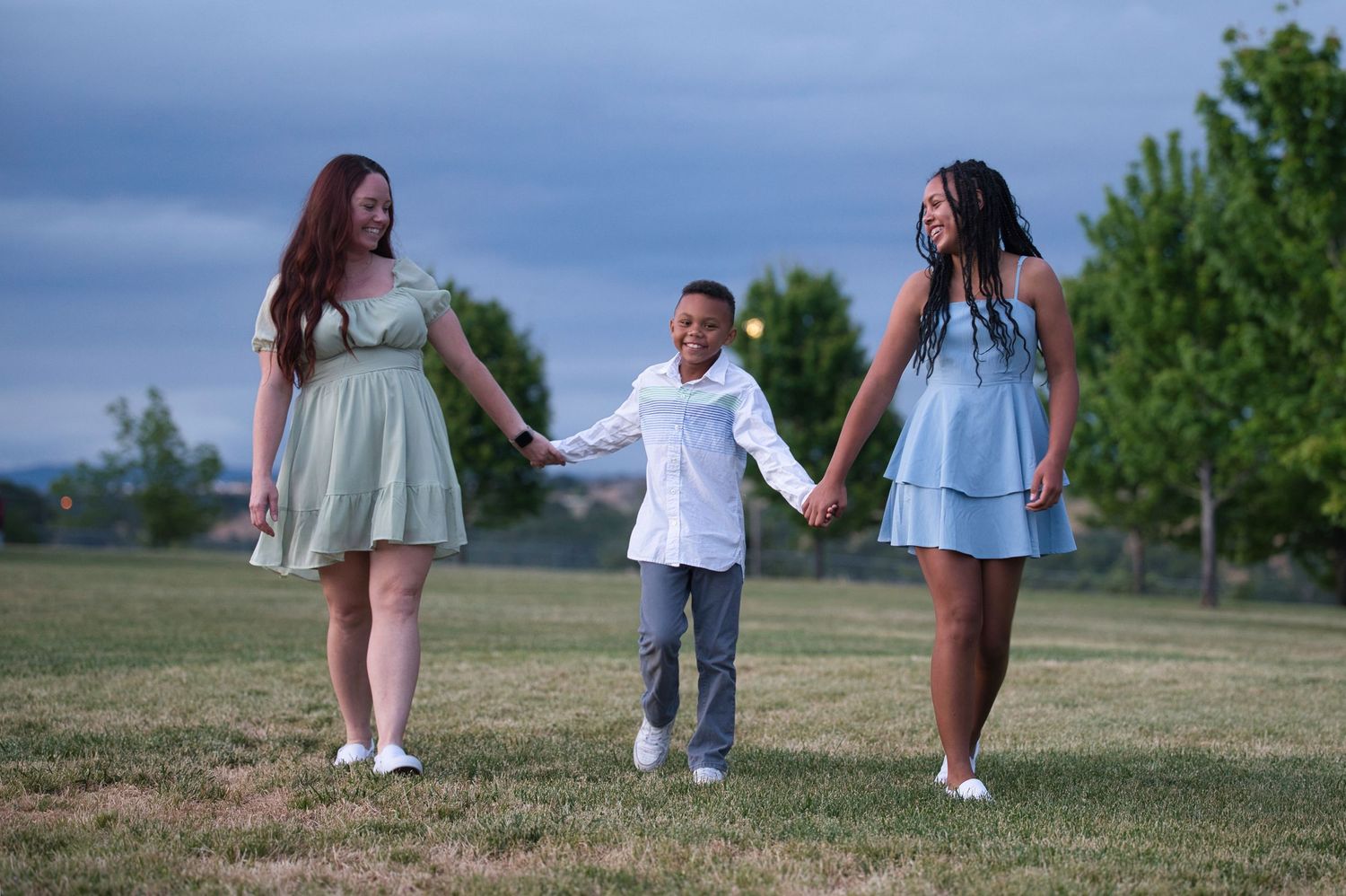 Two women in mint and blue dresses holding hands with a young boy walking in a park.