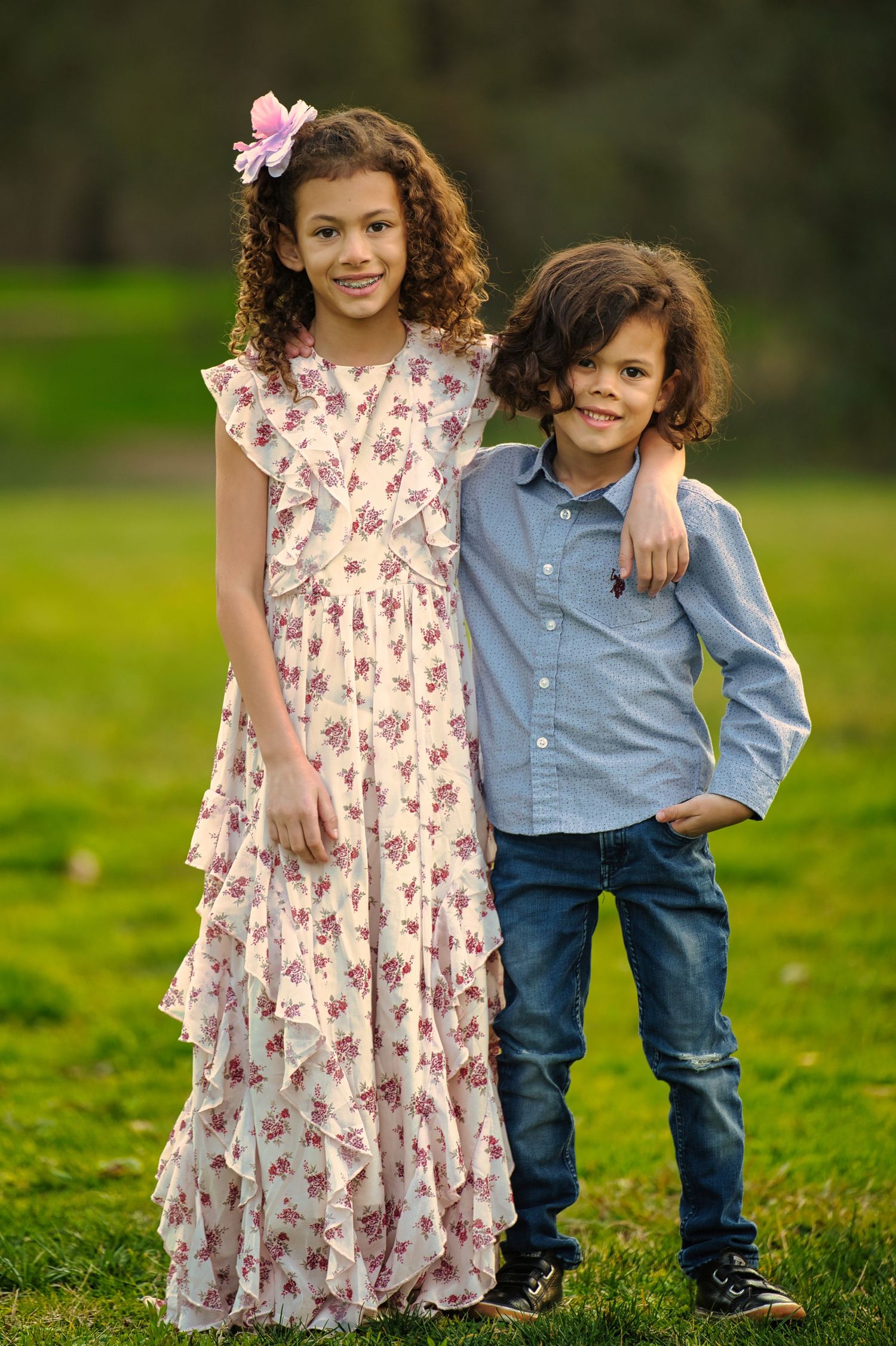 Two curly-haired siblings pose outdoors, girl in floral dress with pink hair bow, boy in blue shirt and jeans.