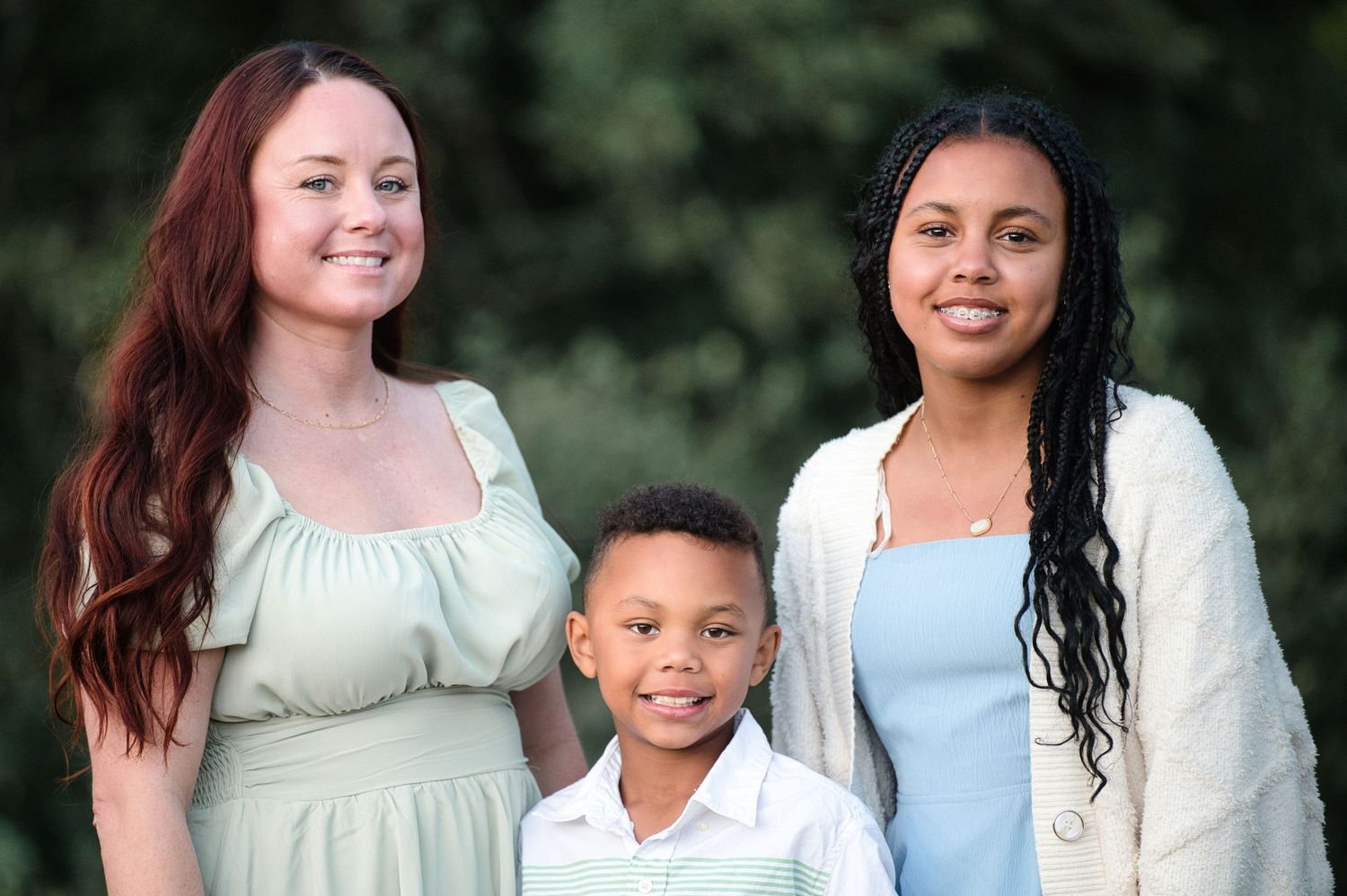 Two women and a young boy smile together outdoors in front of lush green trees for a family portrait.
