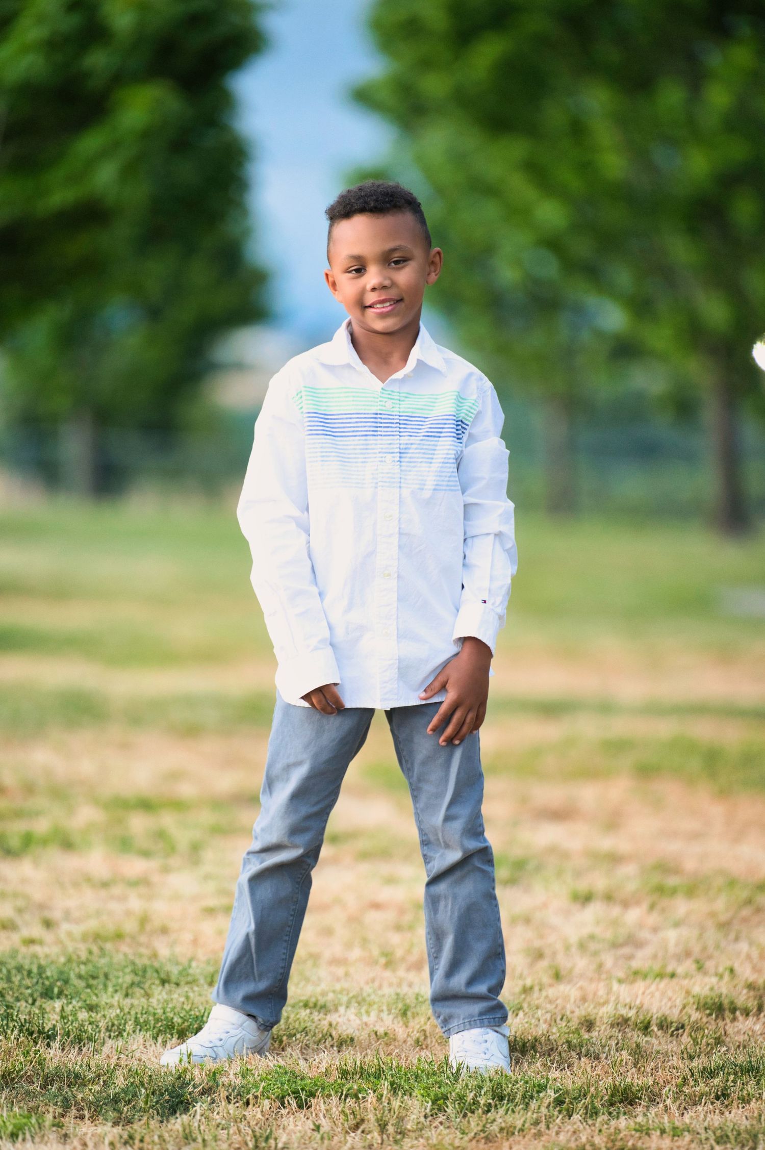 Young boy in white striped shirt and jeans stands confidently on dry grass between rows of green trees.