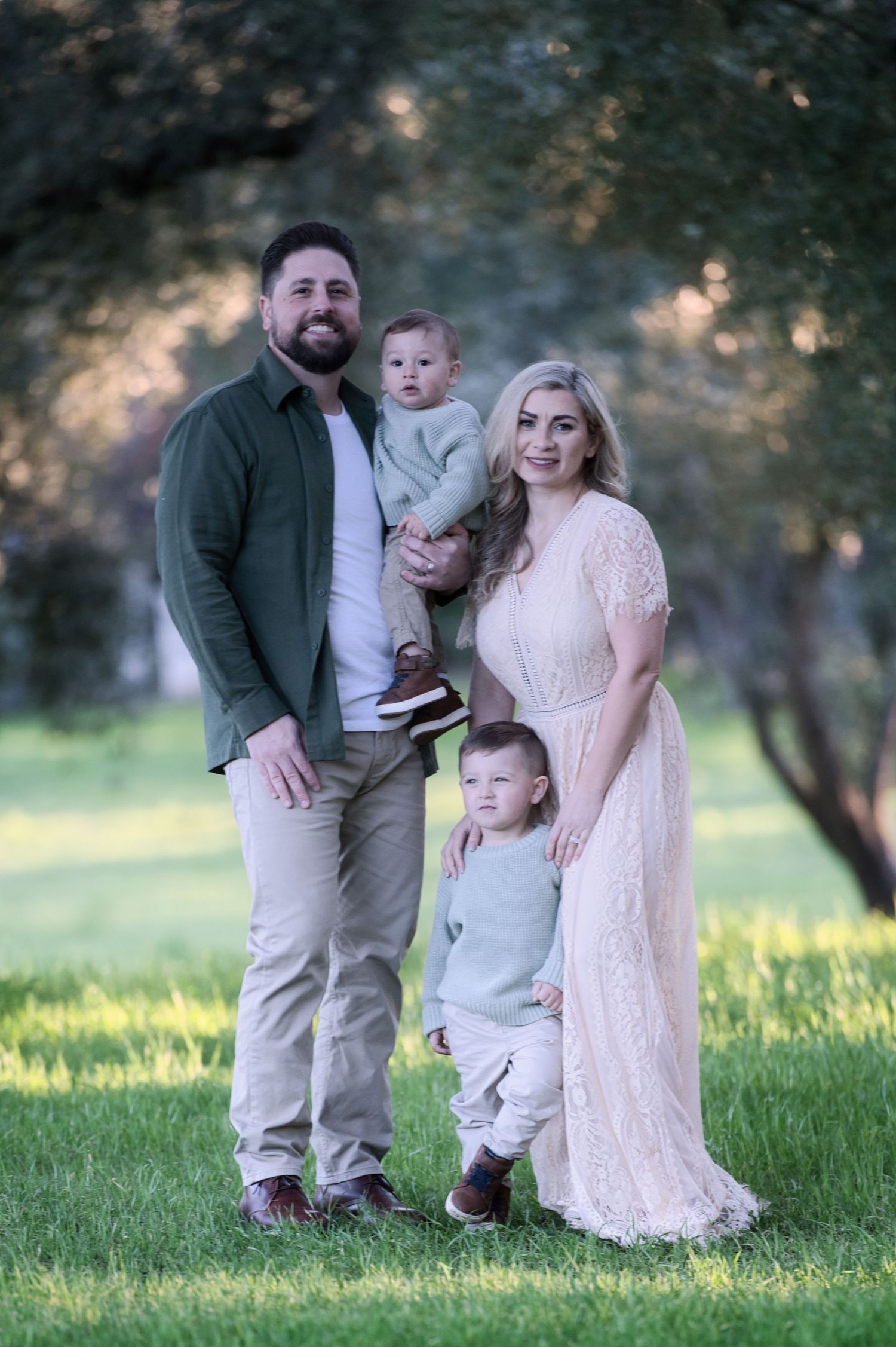 A happy family of four poses in a sunny green park, parents with two young boys.