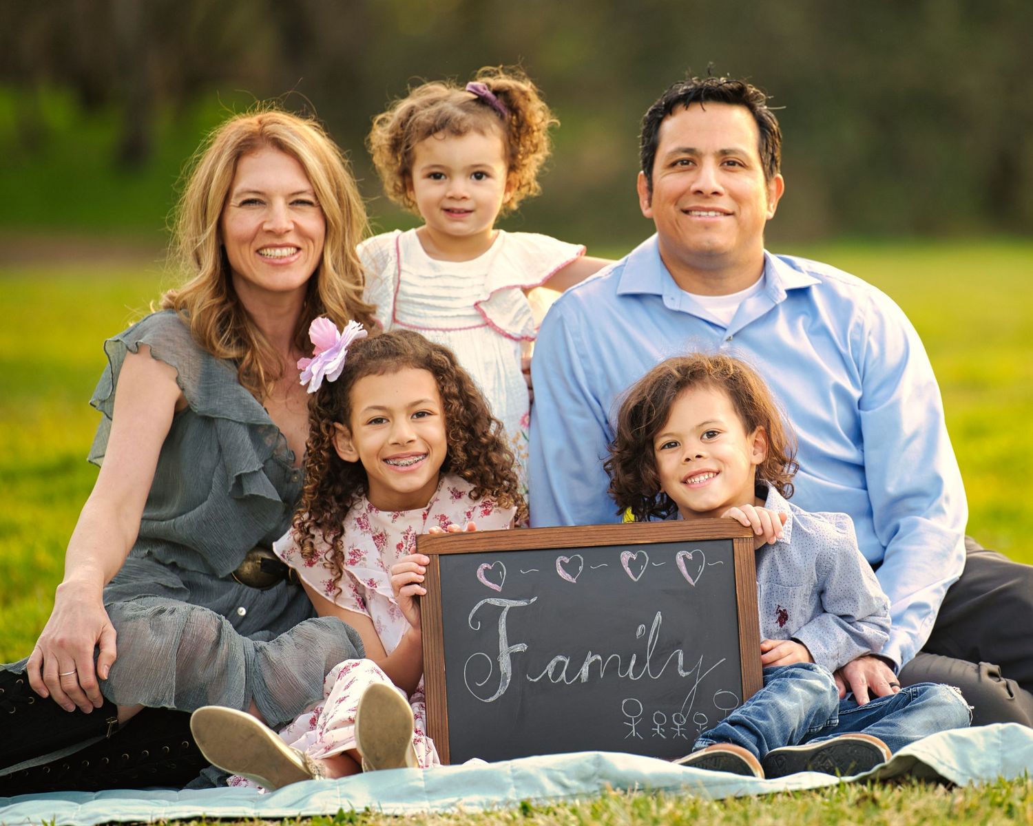 A happy family of five sits on a blanket outdoors holding a chalkboard sign that reads Family.