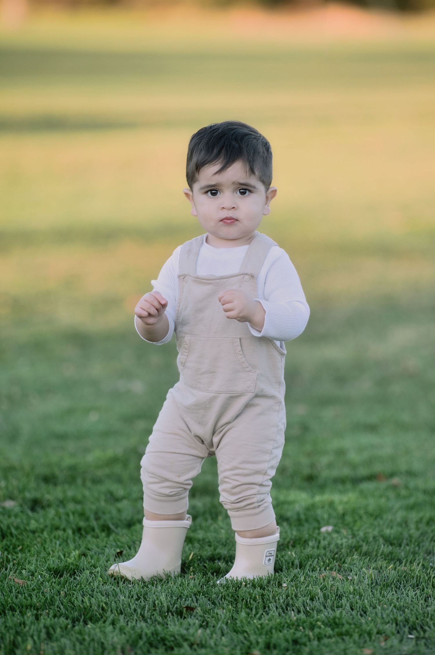 An adorable baby in beige overalls stands barefoot on a green grassy lawn.