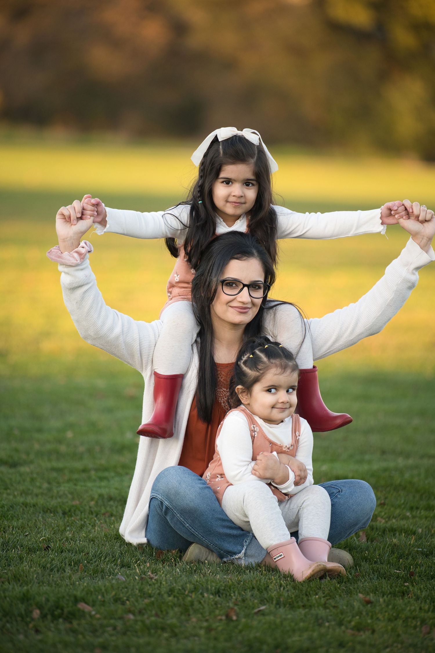 Smiling mother sitting on grass with two little girls raising their arms in a golden autumn park.