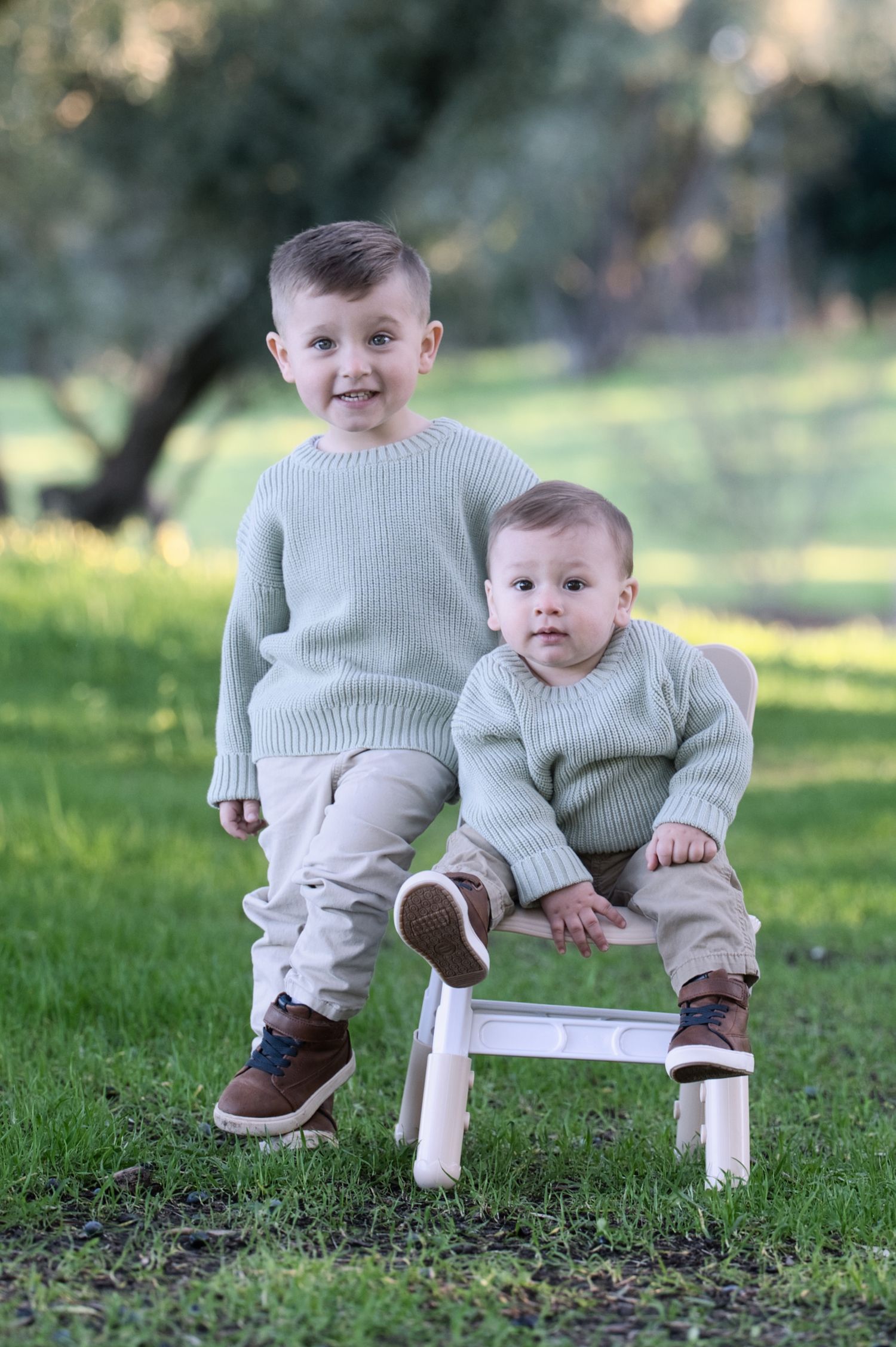 Two brothers in matching sage green knit sweaters posing outdoors, older boy standing beside toddler on white chair.