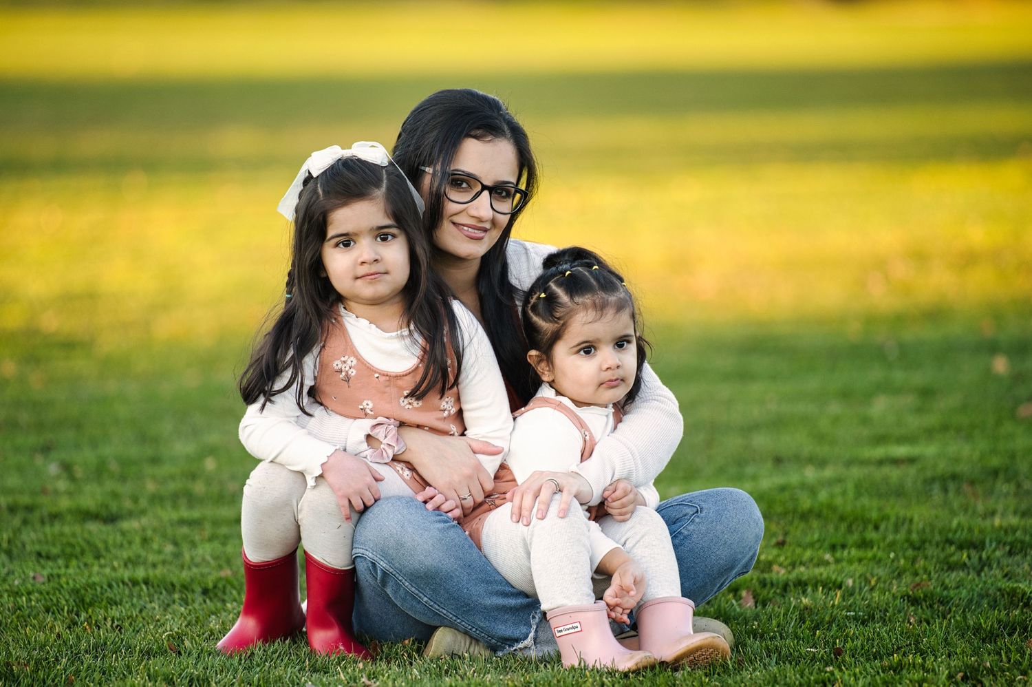 Smiling mother with glasses sitting on grass hugging two young daughters in park.