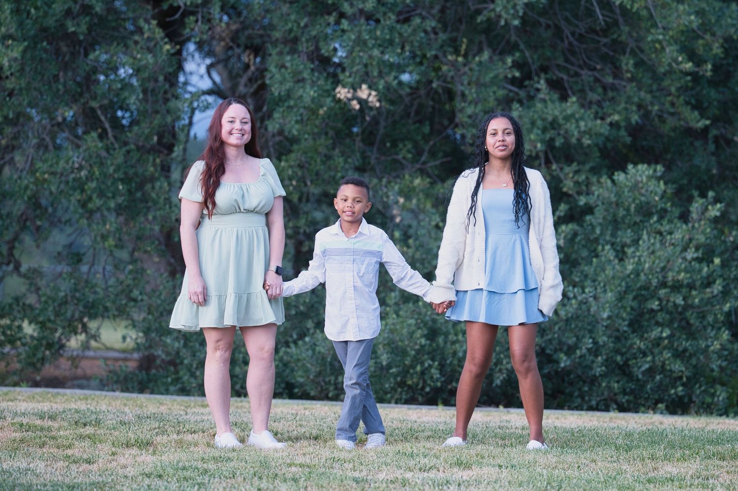 Two girls in pastel dresses hold hands with a young boy while walking together on a grassy park lawn.