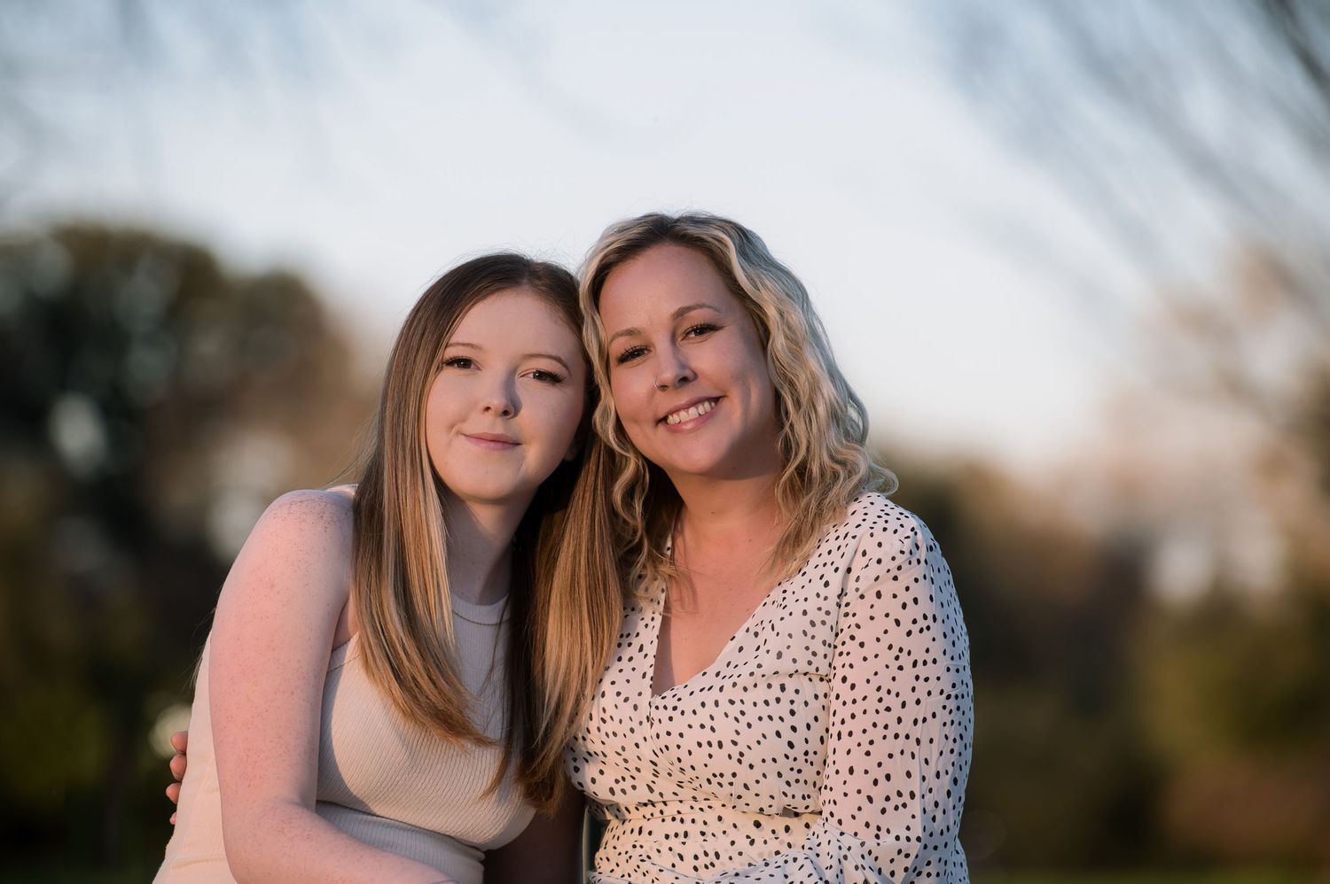 A mother and daughter lean close together smiling warmly outdoors in soft evening light.