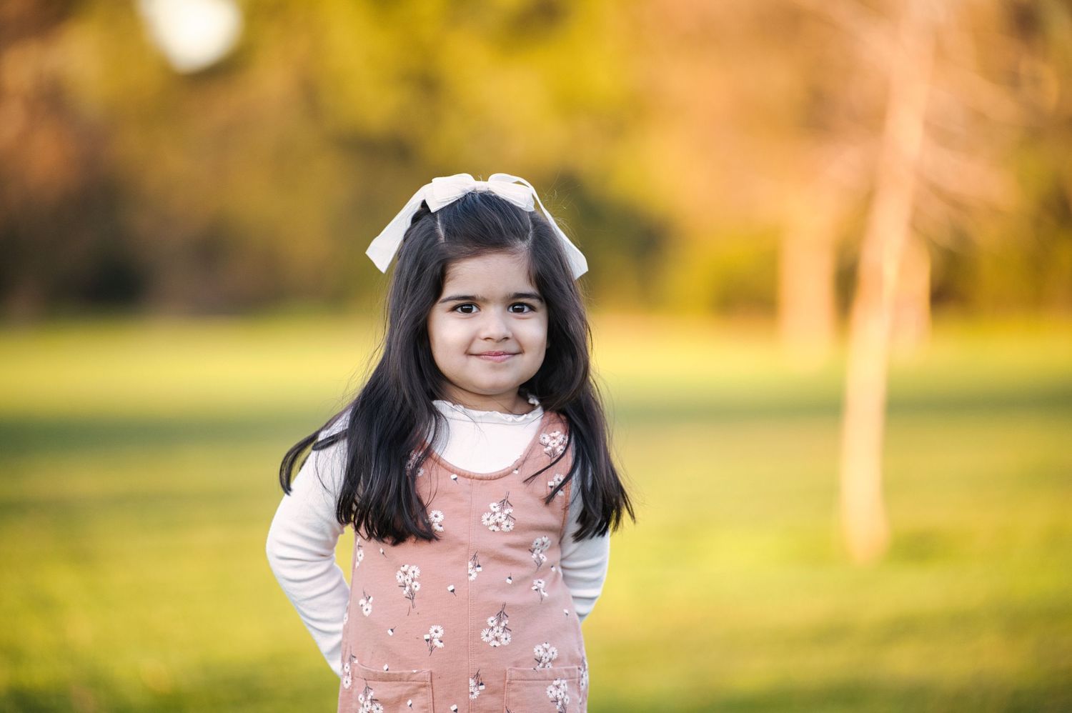Young girl in pink floral dress with white bow smiling in sunny park setting.