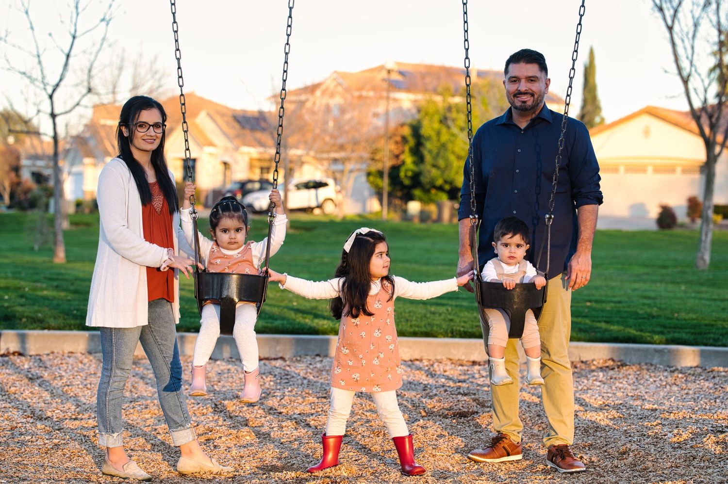 A family of five poses at a playground with two children on swings and a toddler standing between the parents.