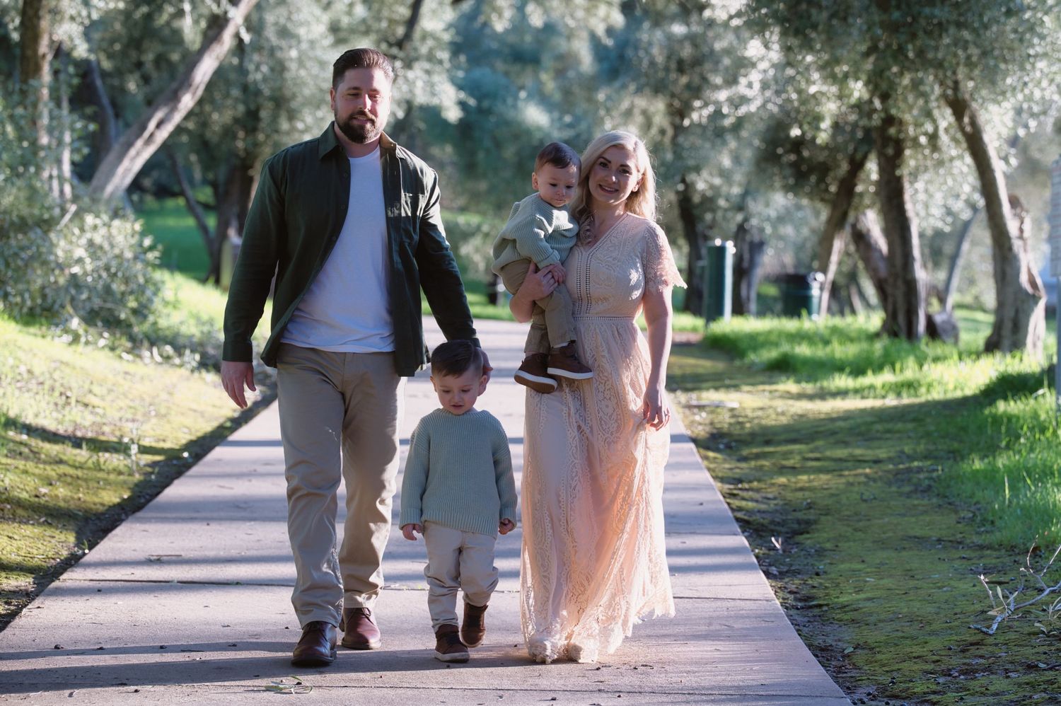 Family of four walking along a tree-lined path, mother in flowy dress carrying baby while toddler walks ahead.