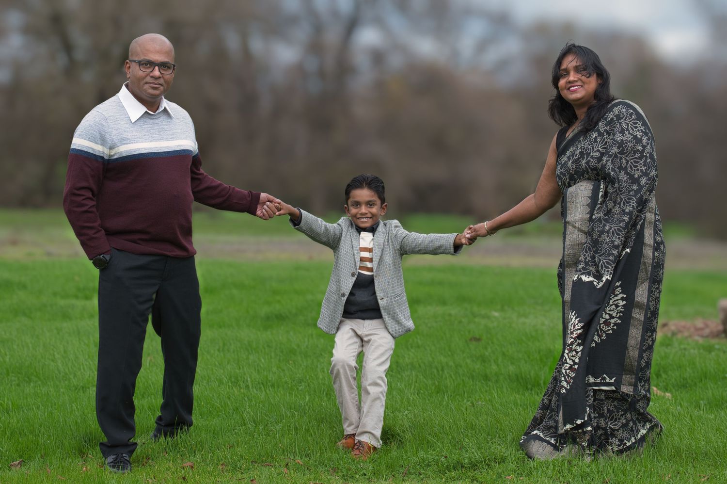 Family of three holding hands in a green field, parents swinging young boy between them.