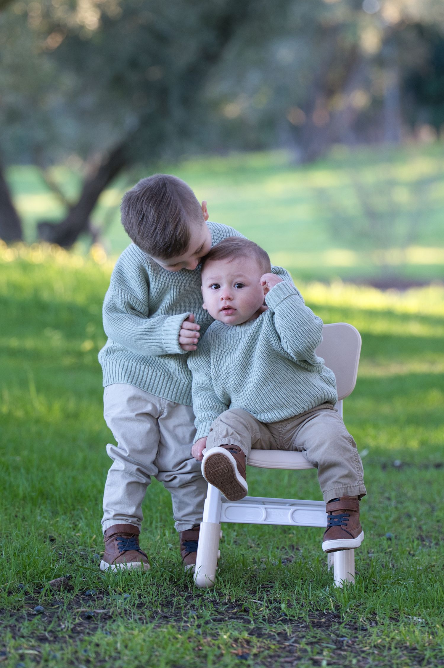 Sweet toddler boy kissing baby sibling seated on small white chair during outdoor photo session in green park.
