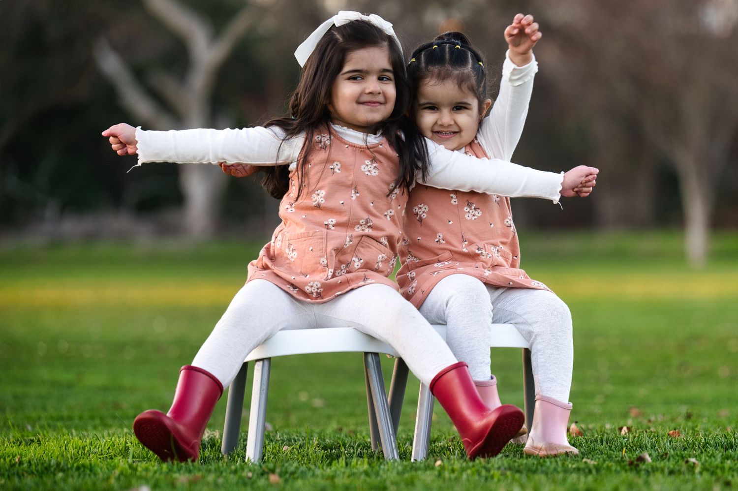 Two young sisters in matching pink dresses and red boots sit back-to-back on a chair in a green park.