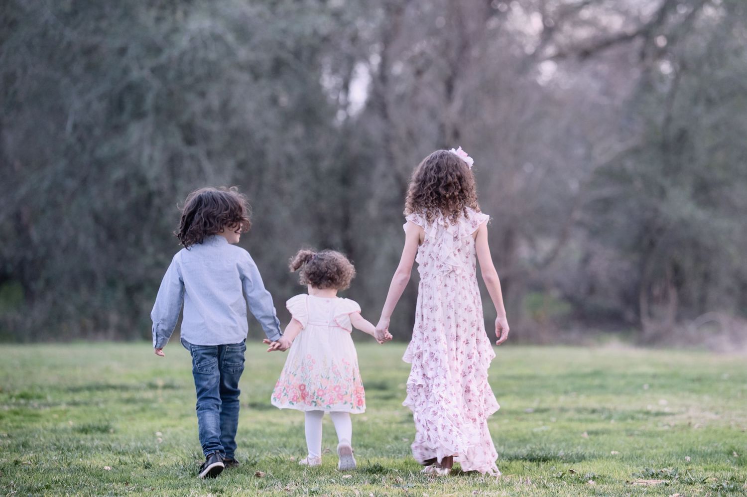 Three young children hold hands while walking through a lush green park from behind.