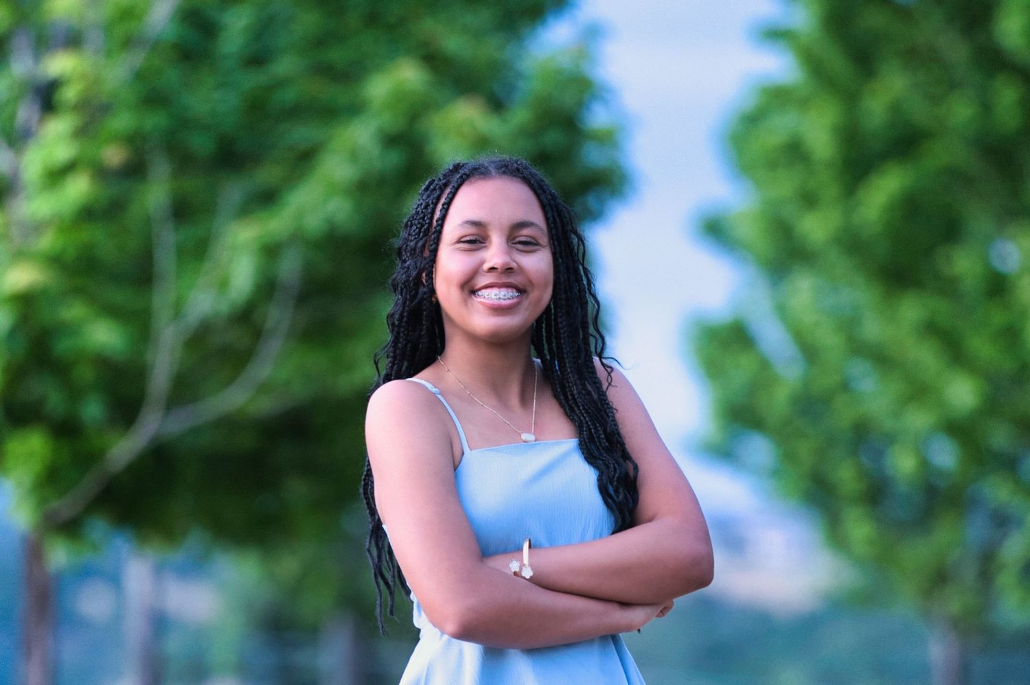 A young woman with long braids in a blue dress stands smiling with arms crossed in a park.