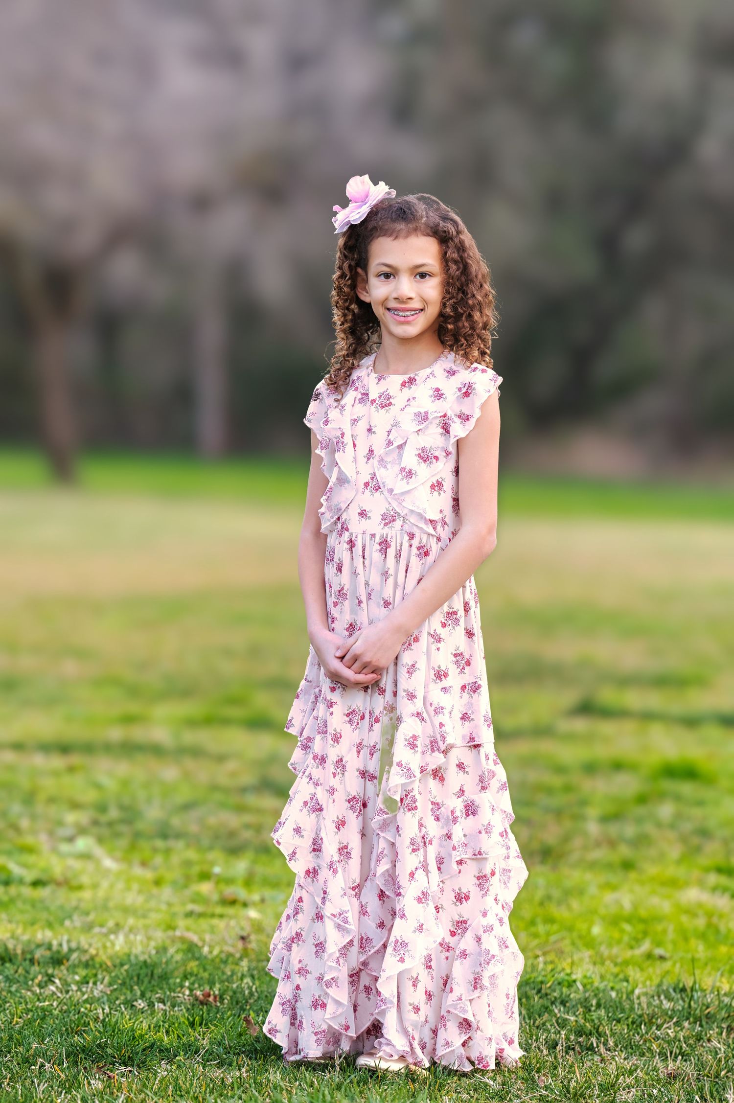 Young girl in floral maxi dress with pink hair bow smiling outdoors in green park.