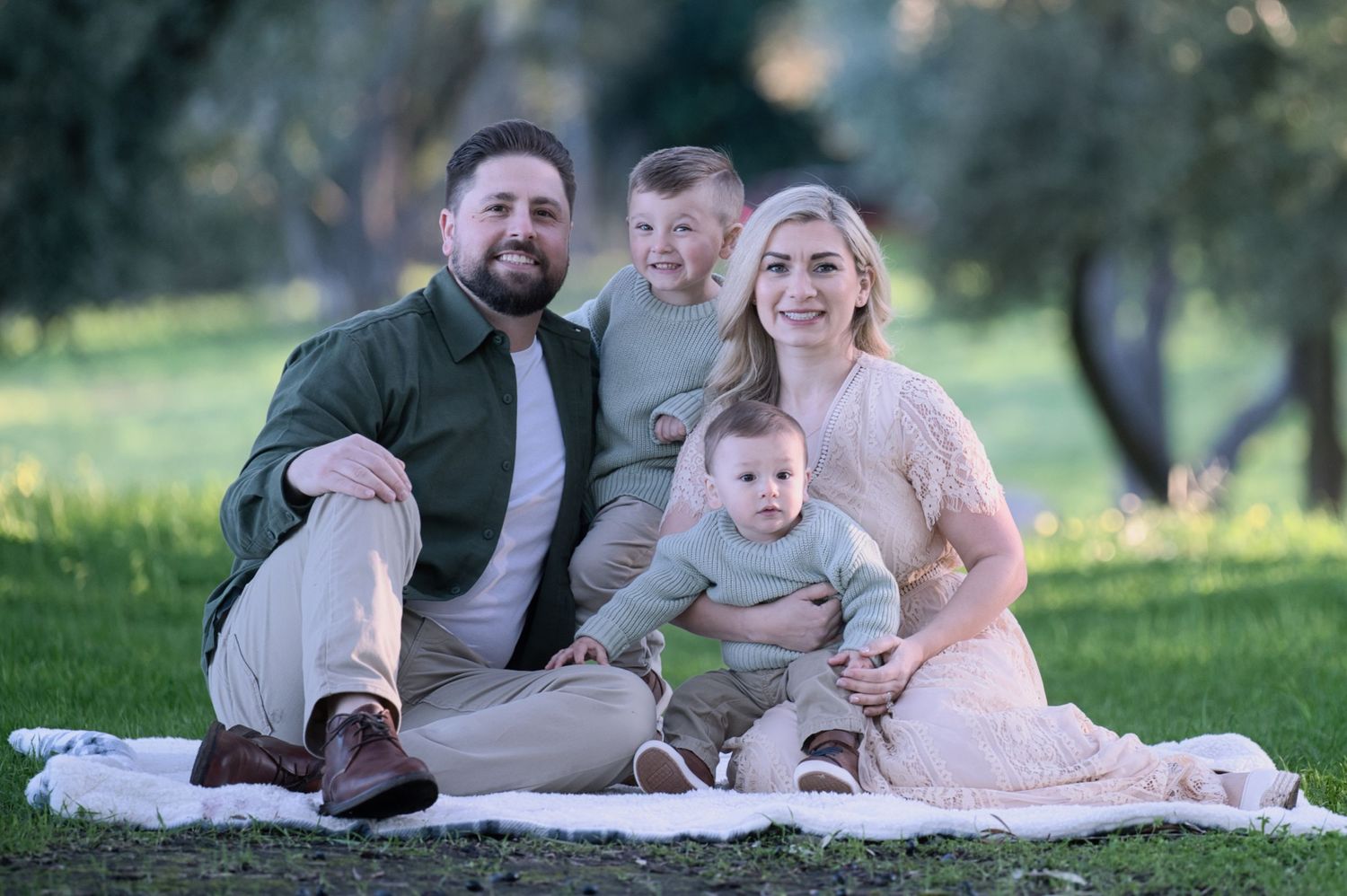 Happy family of four sitting on a blanket in a green park, smiling together on a sunny day.